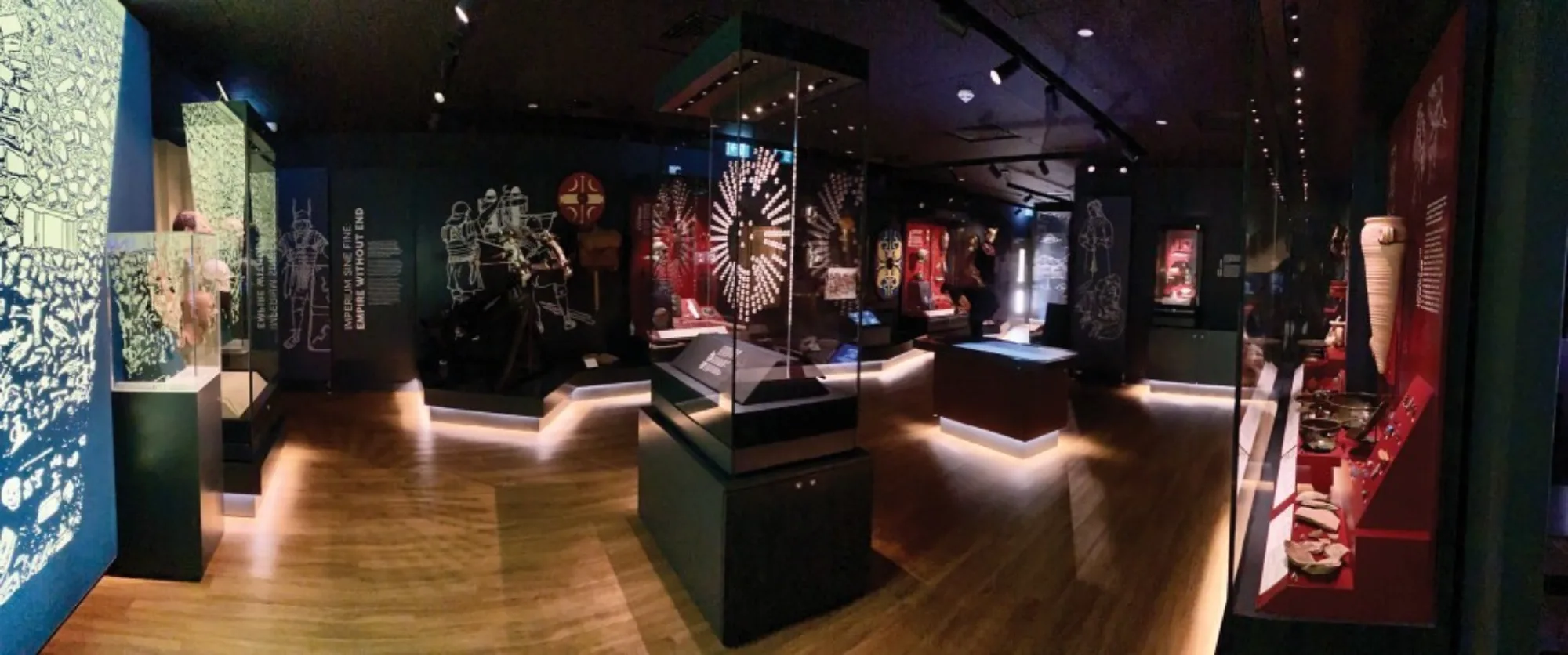 Indoor museum gallery with illuminated display cases containing artifacts, illustrated wall panels, and central glass exhibits, set within a dark room with wooden flooring and spot lighting.