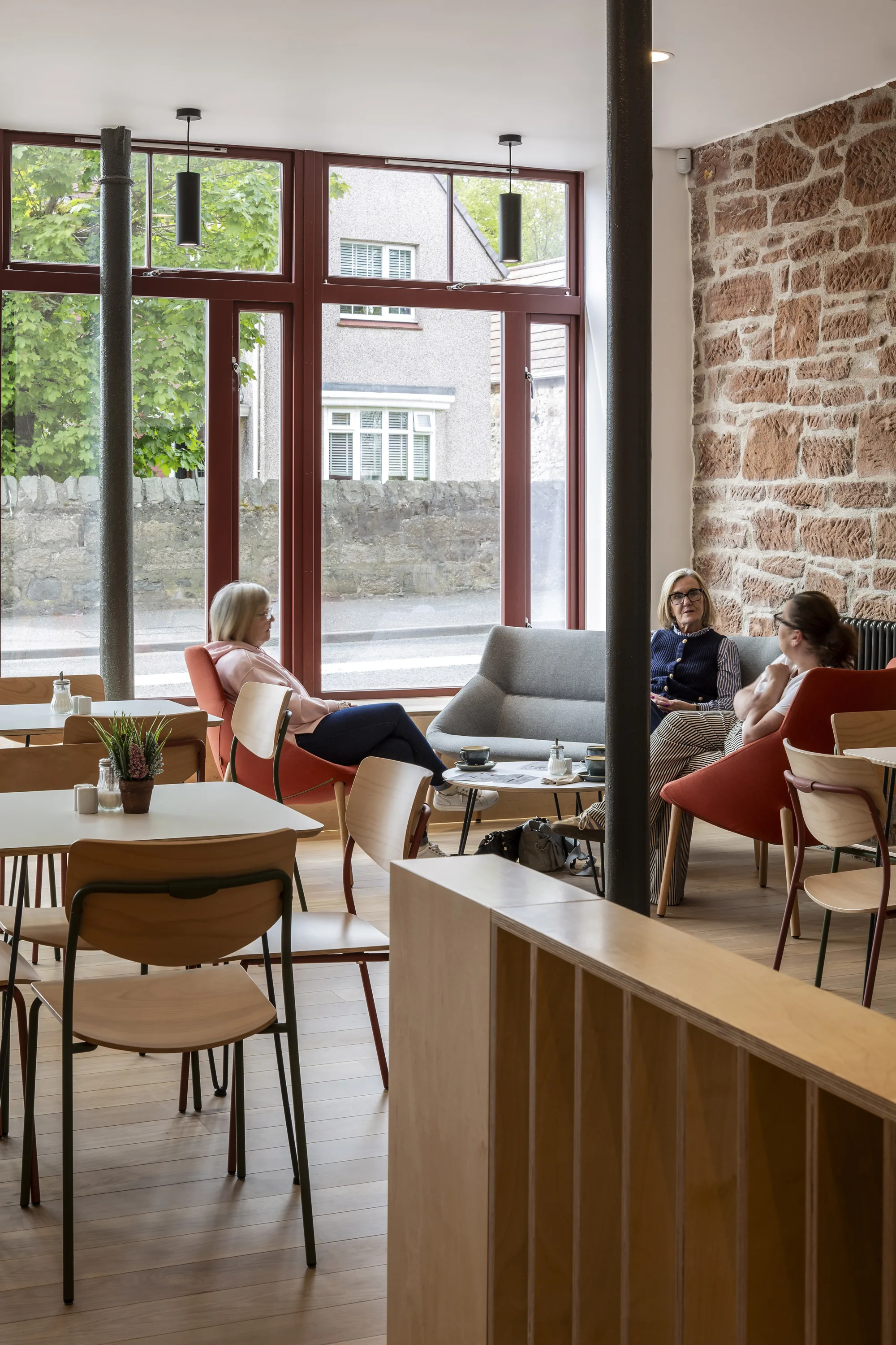 Interior with tables, chairs, large street-facing windows and an exposed stone wall, with several people seated in a lounge area.