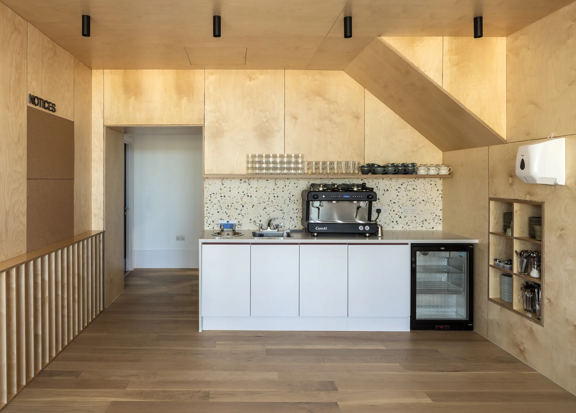 Timber-lined interior with fitted counter, coffee machine, open shelving and wood flooring beneath recessed lighting.