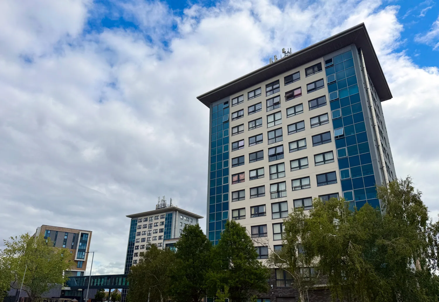 Three modern high-rise buildings with blue and white facades stand against a partly cloudy sky. The tallest is in the foreground; trees and a pedestrian bridge are visible at the base.