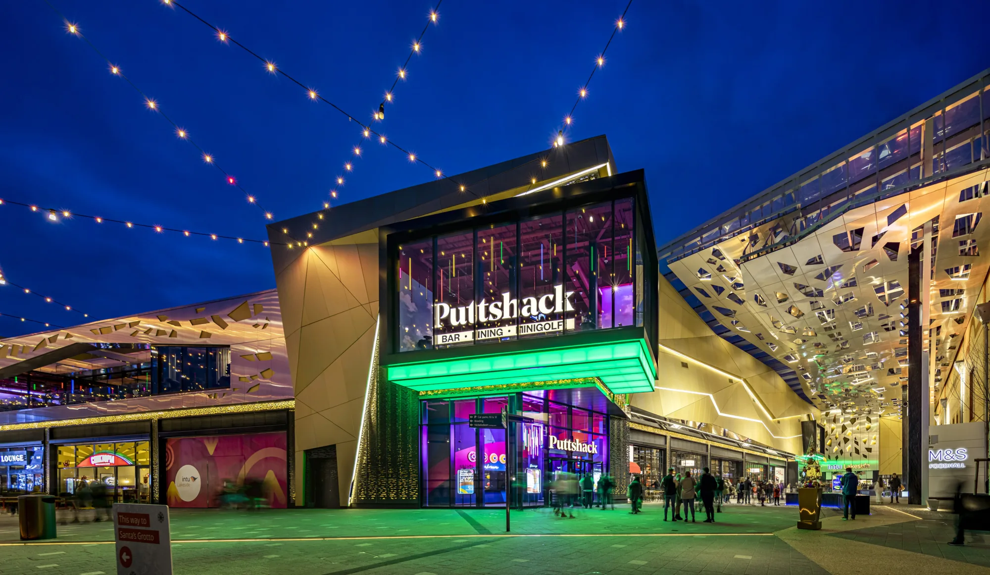 Exterior entrance to Intu Lakeside shopping centre in the evening. With exterior lighting and people shopping