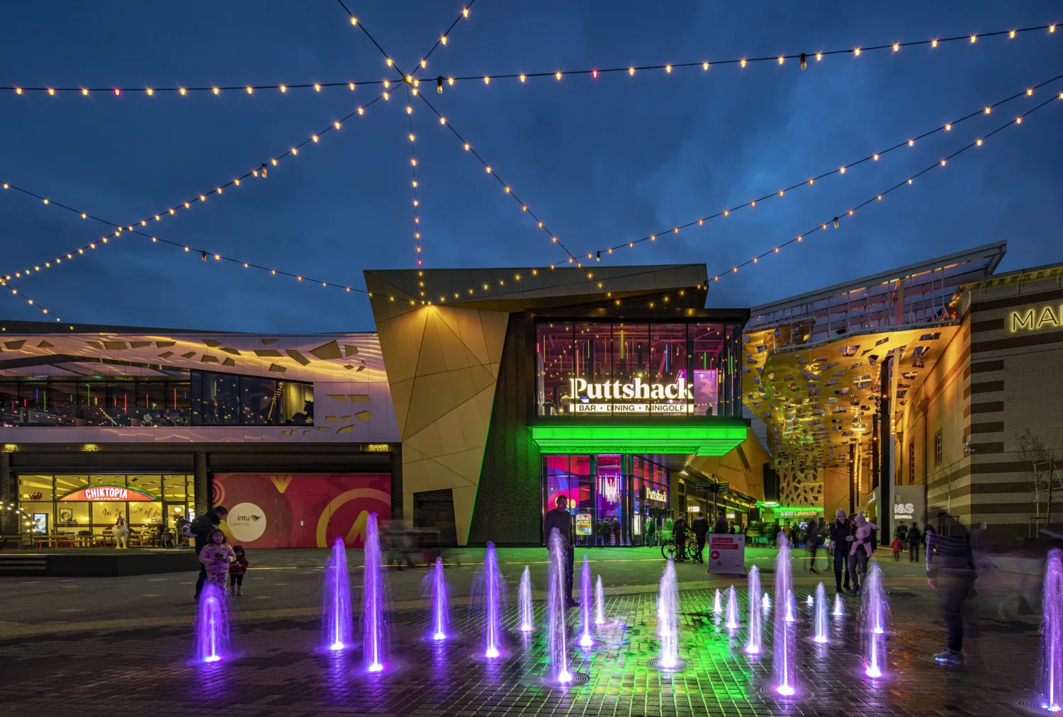 Exterior entrance to Intu Lakeside shopping centre in the evening. With exterior lighting and people shopping