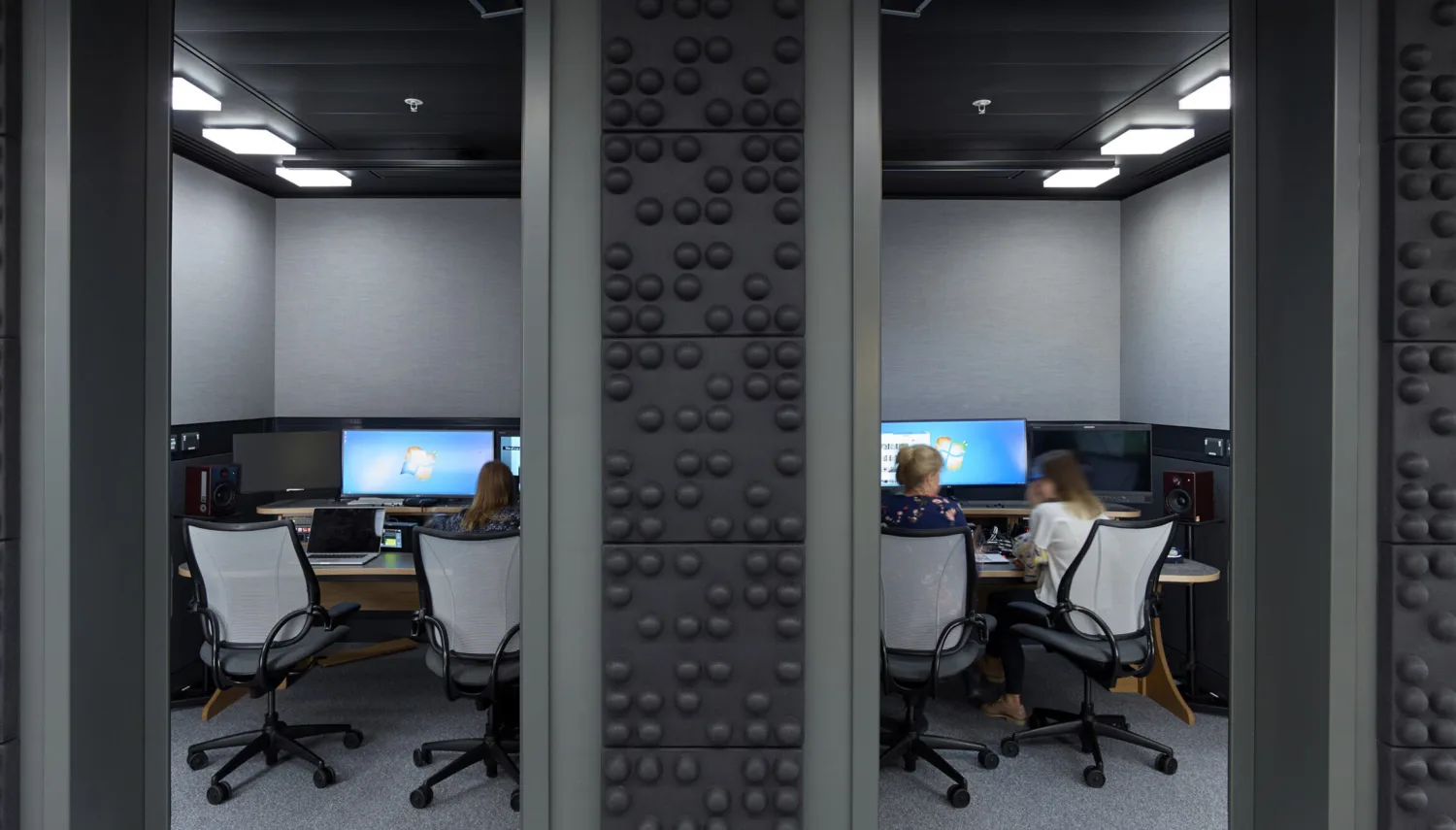 A+E Network meeting rooms with desks, large screens with people working. Grey acoustic walls and grey chairs.