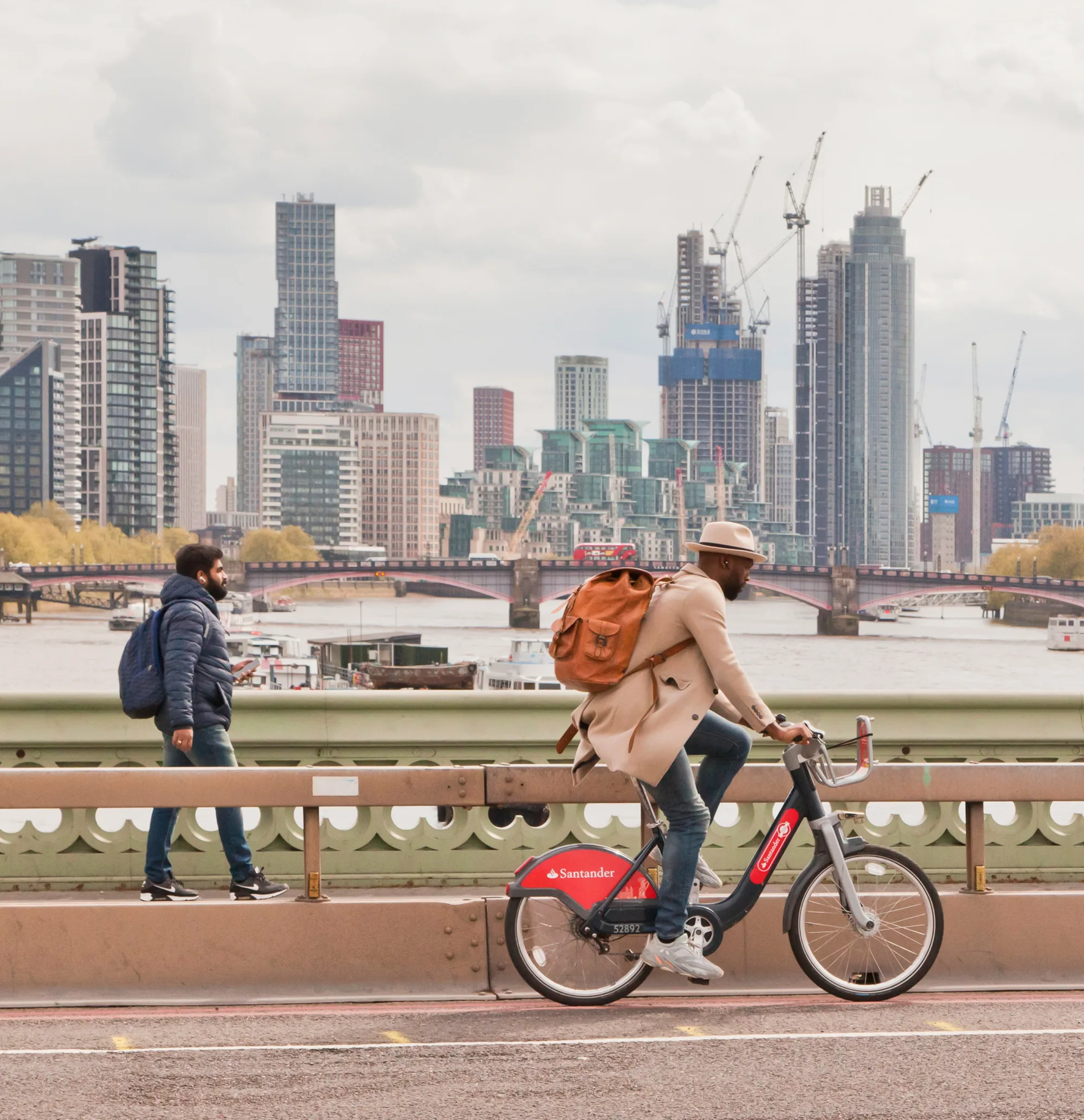 One pedestrian and one cyclist crossing a bridge over the river Thames with high rise buildings under construction in the background