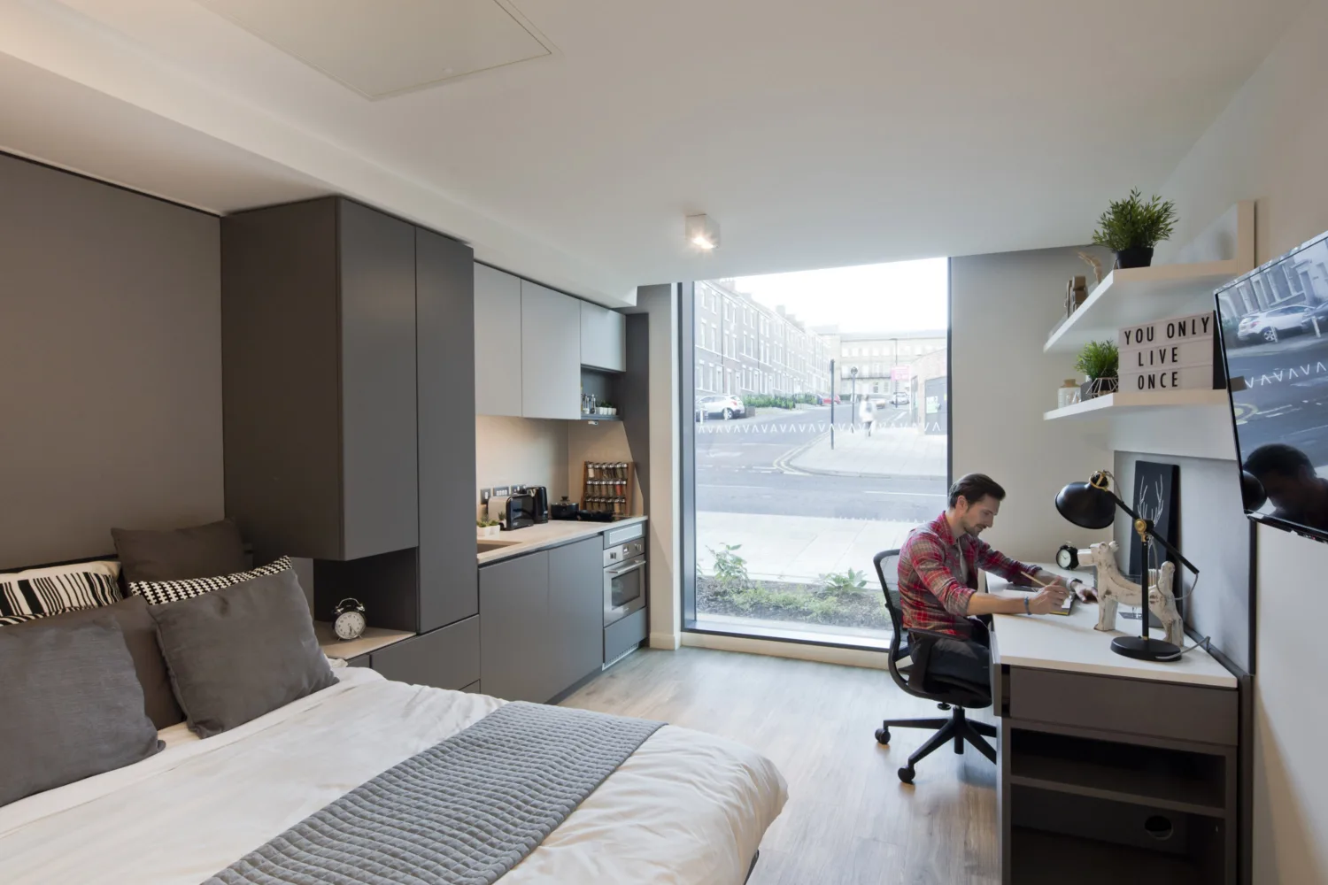 Bedroom in student accommodation with a bed, small kitchen and desk with a large window