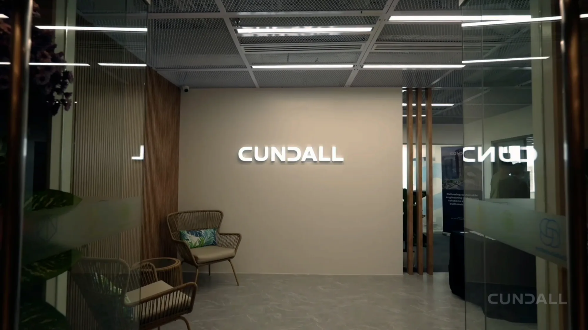 Commercial office interior with illuminated Cundall wall sign, glass partitions, timber slats, seating and ceiling lighting.