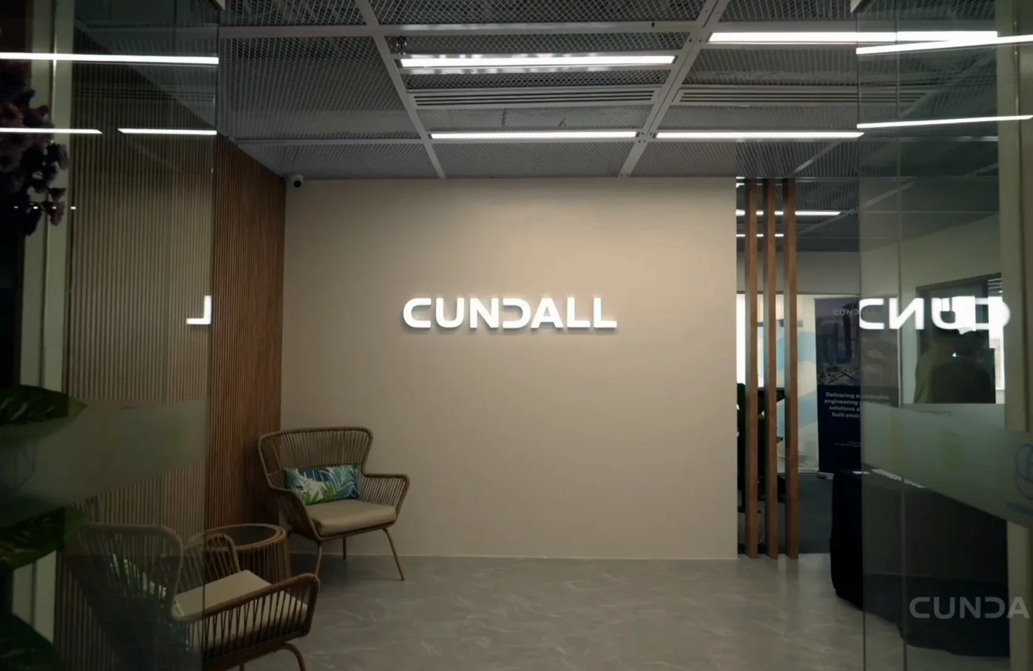 Commercial office interior with illuminated Cundall wall sign, glass partitions, timber slats, seating and ceiling lighting.
