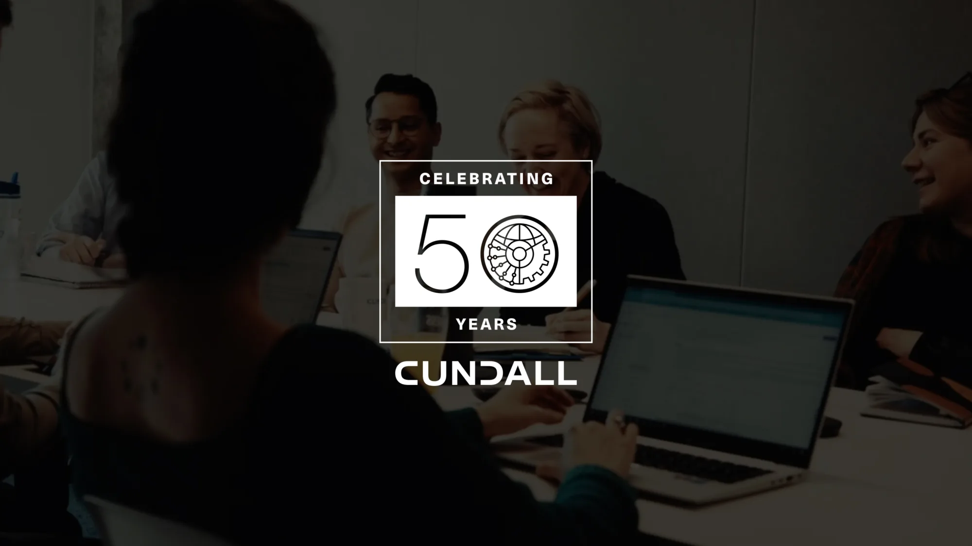 A group of people seated around a meeting table with laptops and notebooks in a modern office setting. Overlaid graphic reads “Celebrating 50 Years Cundall” with a gear and circuit design.