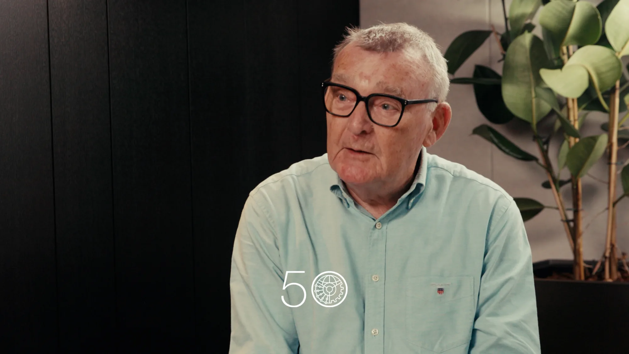A person wearing a light teal button-up shirt sits indoors against a dark wall. A tall plant with broad green leaves stands on the right. A “50” anniversary-style graphic is displayed near the bottom of the image.