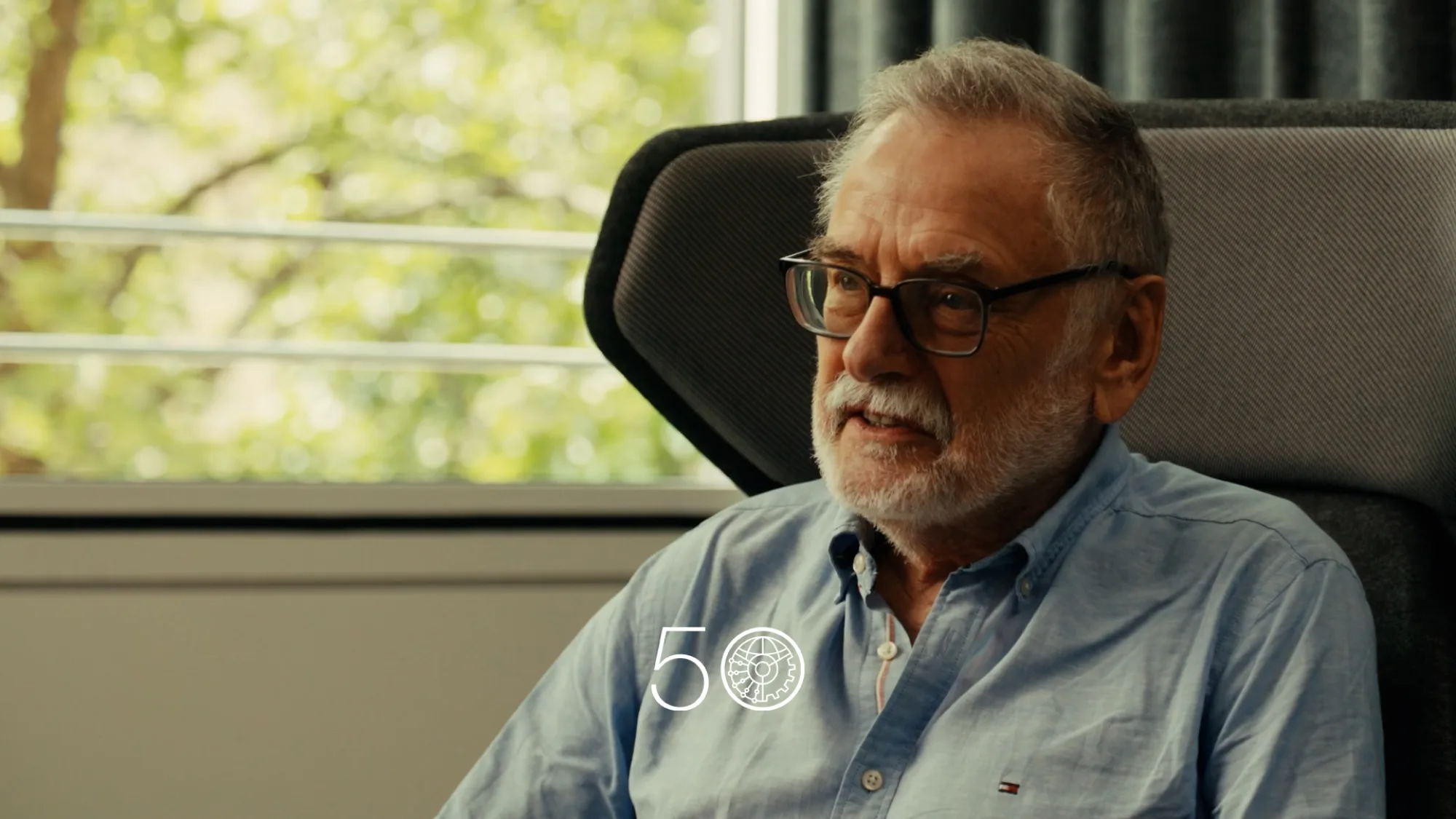 A person in a light blue button-up shirt sits in a modern chair near a large window with green trees visible outside. Dark curtains hang to the right, and a “50” anniversary-style graphic appears at the bottom of the image.