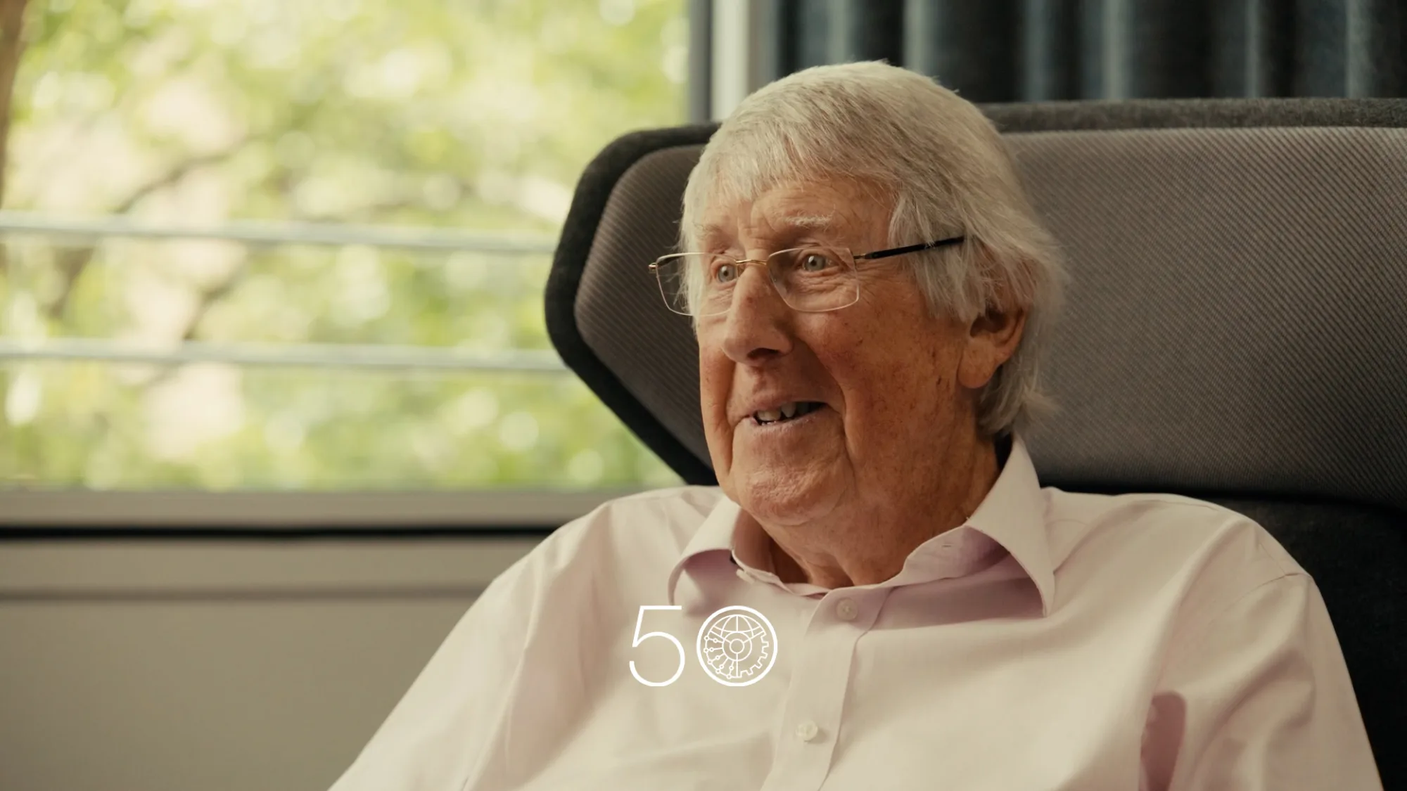 A person wearing a light pink button-up shirt sits in a cushioned chair near a window with green trees visible outside. Dark curtains frame part of the background, and a “50” anniversary-style graphic appears at the bottom of the image.