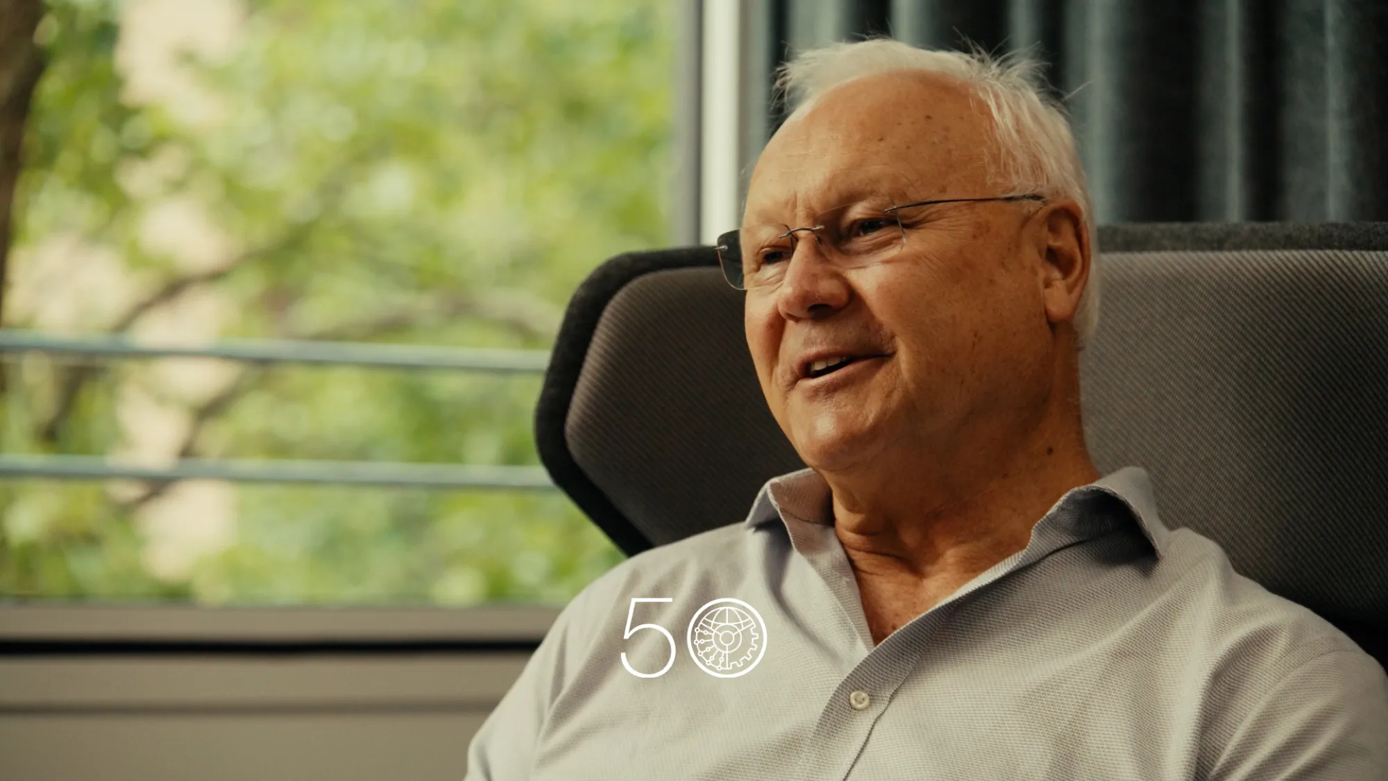 A person in a light grey button-up shirt sits in a cushioned chair near a large window with green trees visible outside. Dark curtains frame part of the background, and a “50” anniversary-style graphic appears at the bottom of the image.