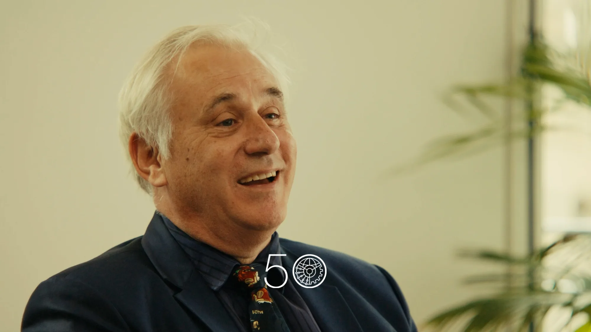 A person wearing a dark suit jacket, navy shirt, and patterned tie sits indoors against a light wall. Green plants frame the right side of the scene, and a “50” anniversary-style graphic appears at the bottom of the image.