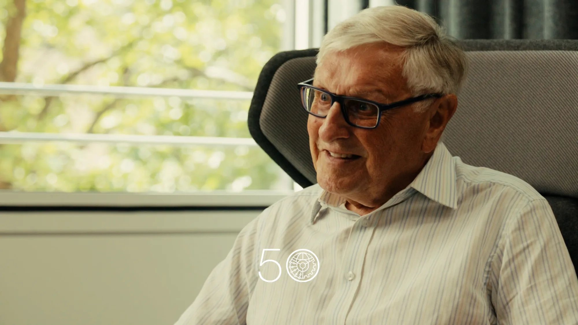 A person wearing a light striped button-up shirt sits in a cushioned chair beside a large window with green trees visible outside. Dark curtains frame part of the background, and a “50” anniversary-style graphic appears at the bottom of the image.