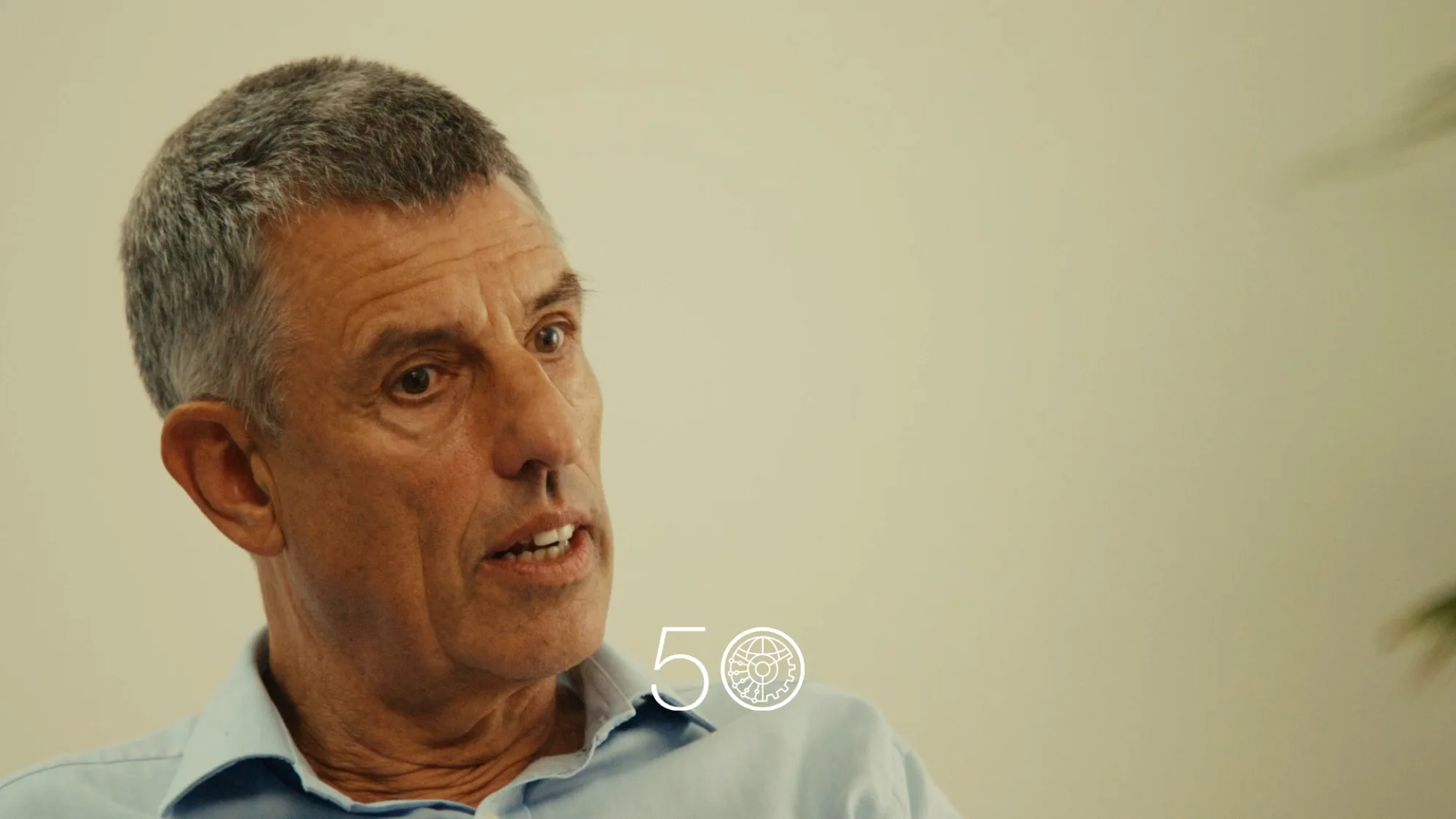 A person in a light blue collared shirt sits indoors against a plain light-colored wall. Part of a green plant is visible on the right, and a “50” anniversary-style graphic appears near the bottom of the image.