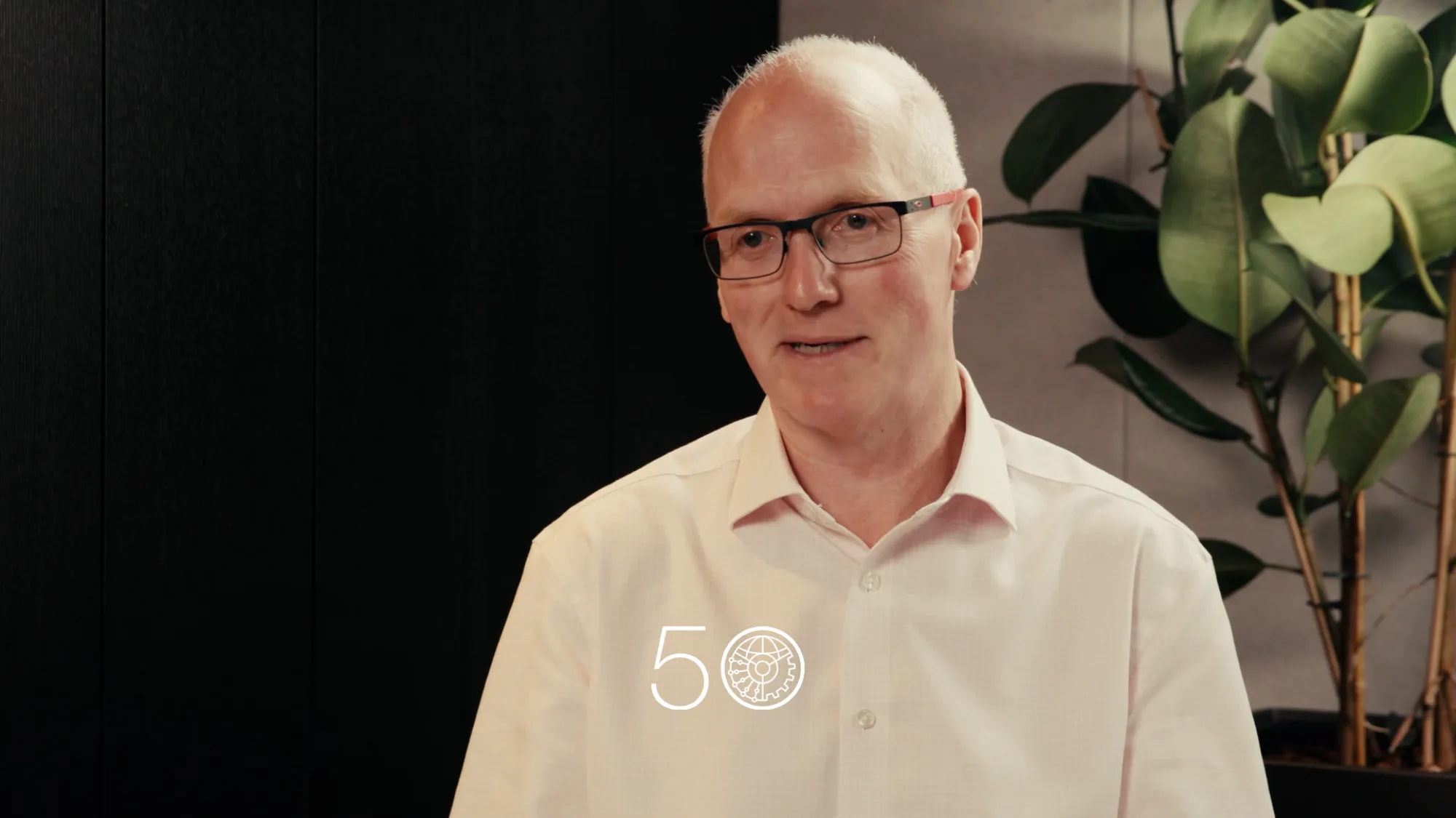 A person wearing a light pink button-up shirt sits indoors against a dark wall, with a tall green plant on the right. A “50” anniversary-style graphic is displayed near the bottom of the image.