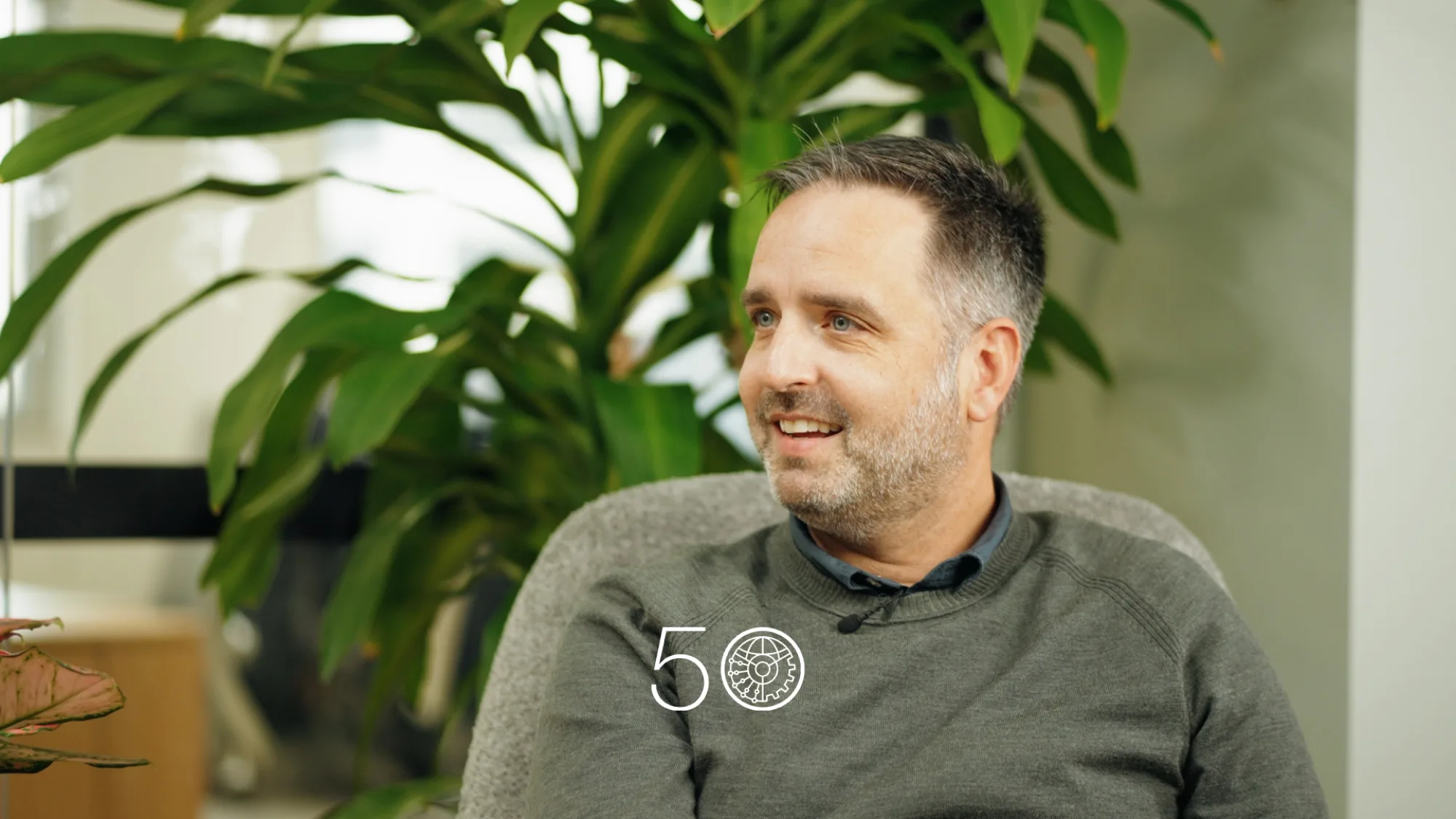 A person wearing a grey sweater sits in an indoor space with large green plants behind them. Part of a window and office interior is visible, and a “50” anniversary-style graphic appears near the bottom of the image.