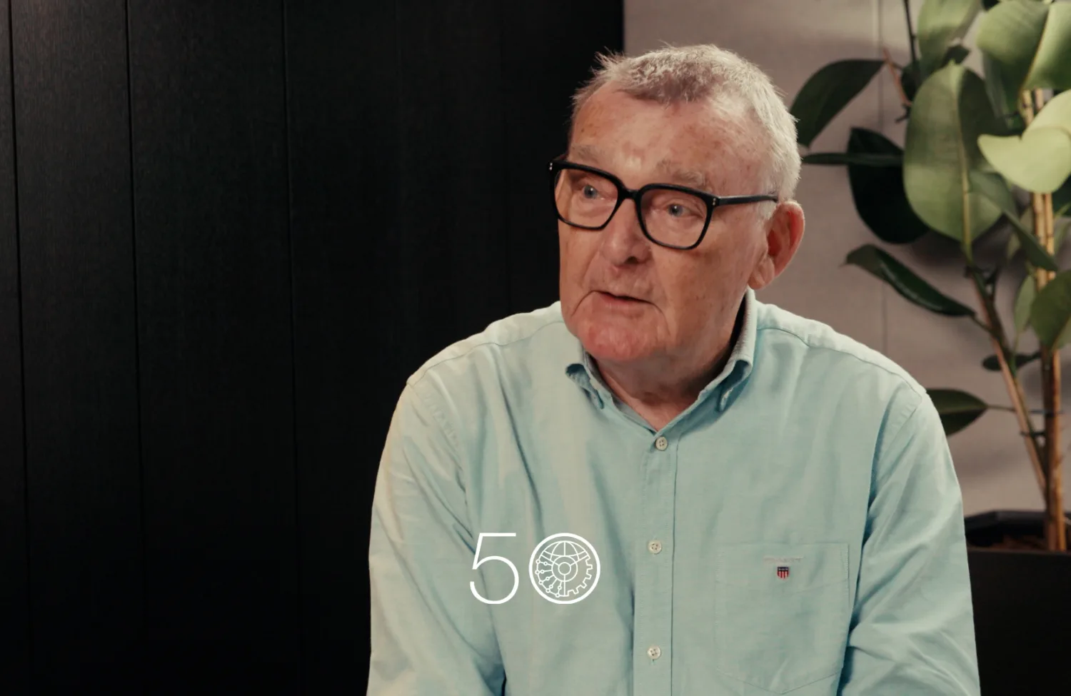 A person wearing a light teal button-up shirt sits indoors against a dark wall. A tall plant with broad green leaves stands on the right. A “50” anniversary-style graphic is displayed near the bottom of the image.