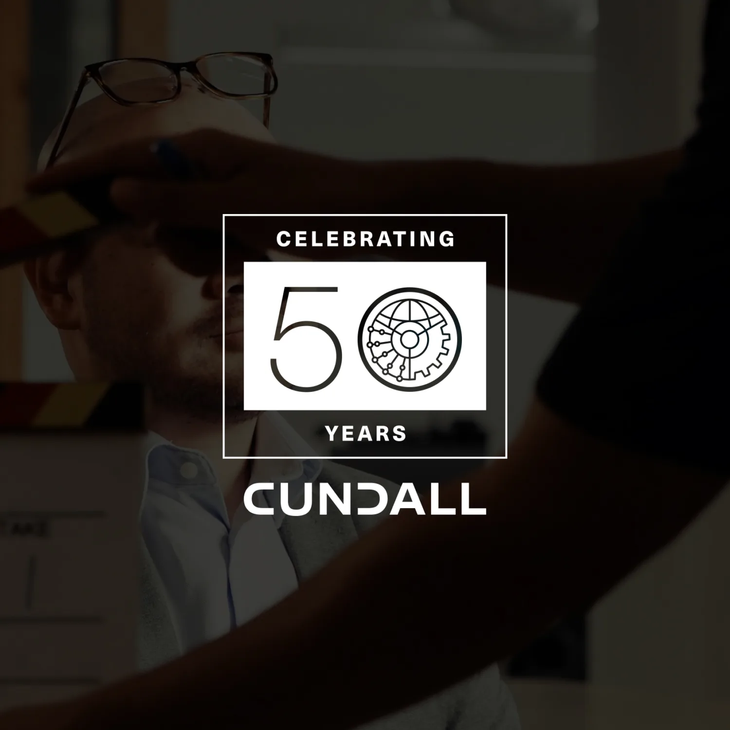 A person seated indoors with glasses resting on their head, while another person holds a clapperboard in front of them. Overlaid text reads “Celebrating 50 Years Cundall” with a gear icon in the design.