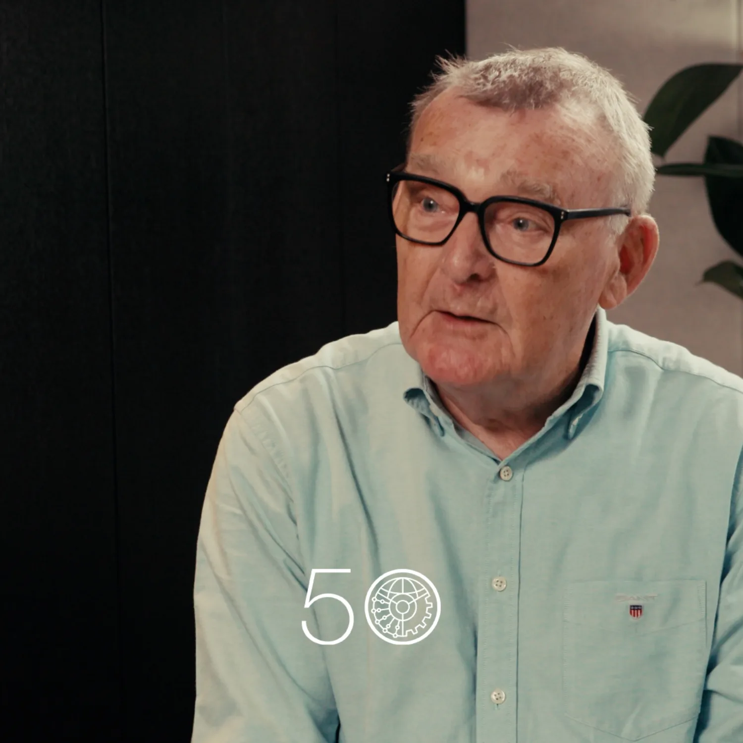 A person wearing a light teal button-up shirt sits indoors against a dark wall. A tall plant with broad green leaves stands on the right. A “50” anniversary-style graphic is displayed near the bottom of the image.
