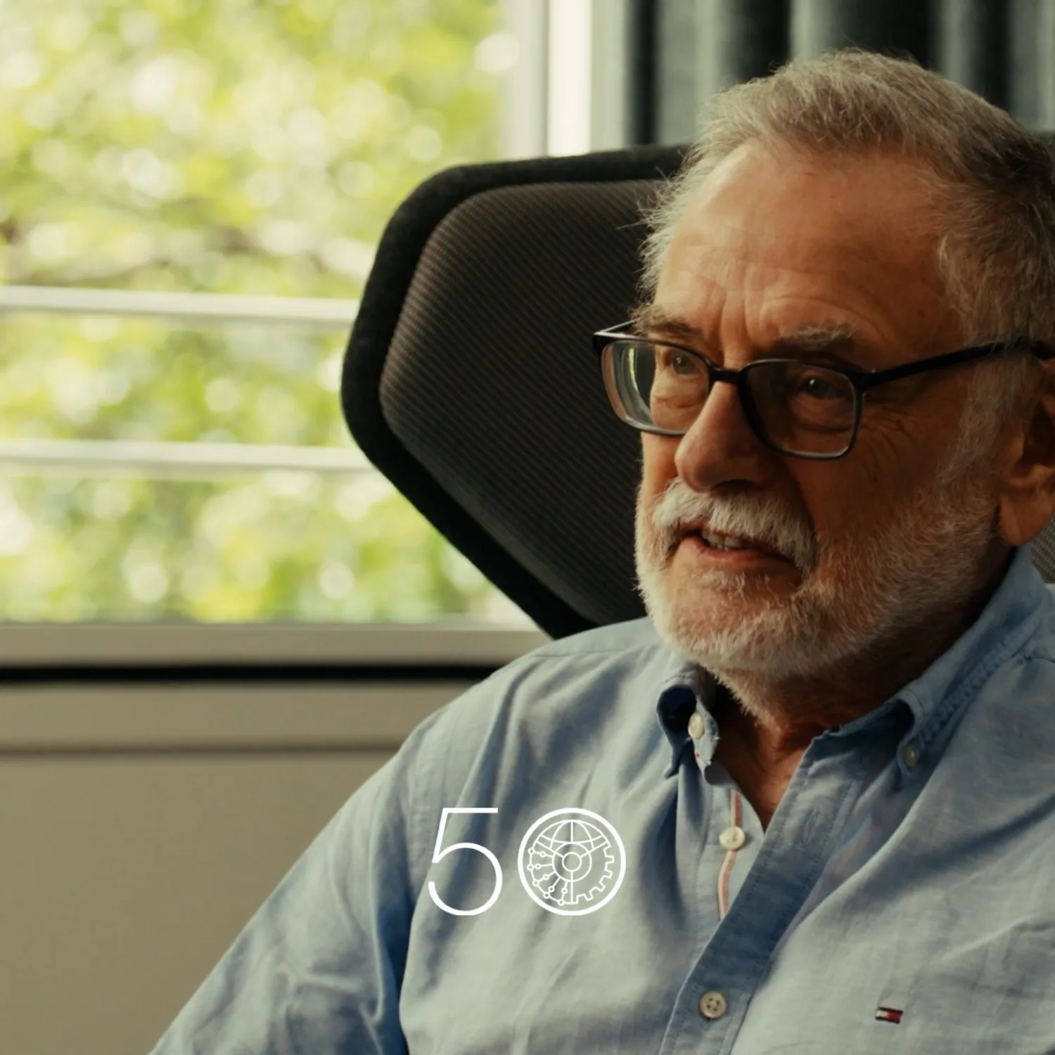 A person in a light blue button-up shirt sits in a modern chair near a large window with green trees visible outside. Dark curtains hang to the right, and a “50” anniversary-style graphic appears at the bottom of the image.