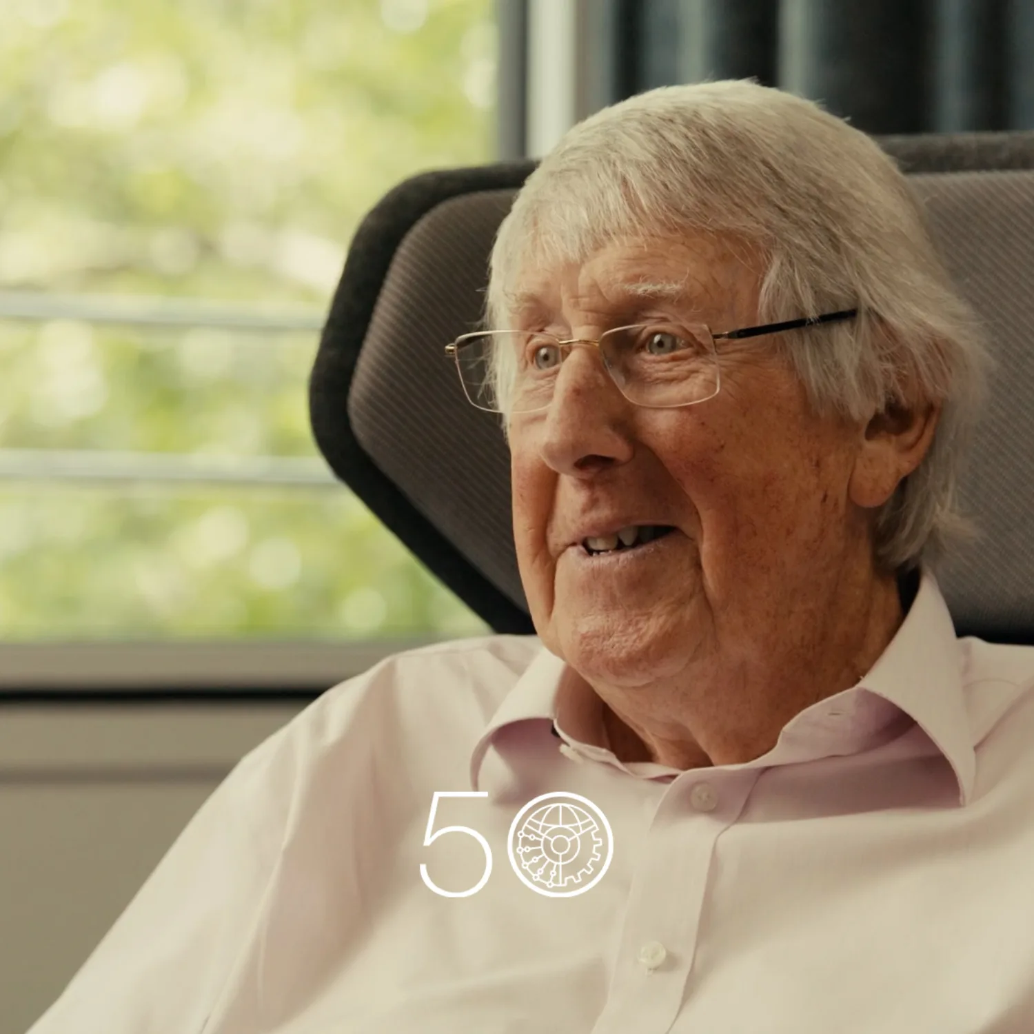 A person wearing a light pink button-up shirt sits in a cushioned chair near a window with green trees visible outside. Dark curtains frame part of the background, and a “50” anniversary-style graphic appears at the bottom of the image.