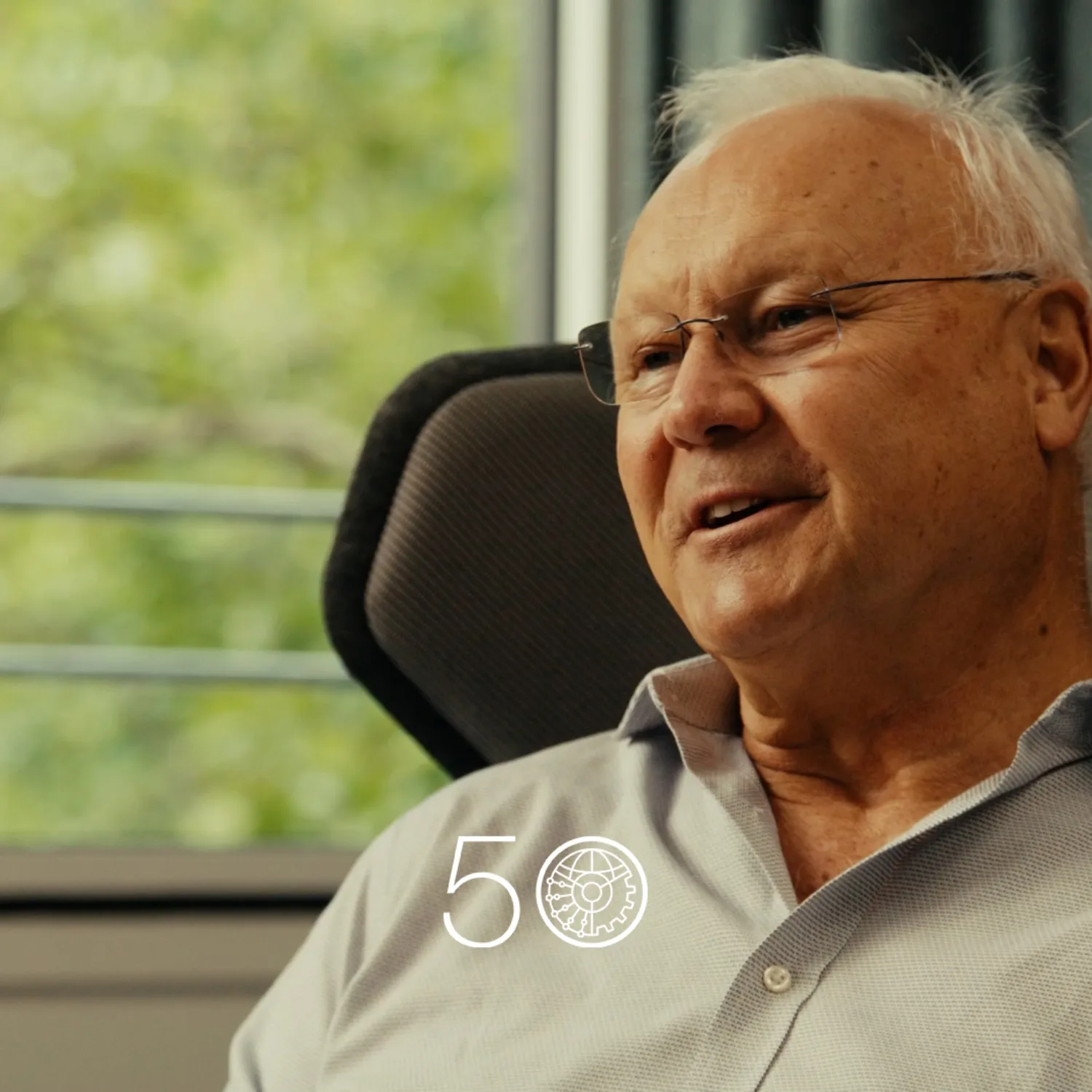 A person in a light grey button-up shirt sits in a cushioned chair near a large window with green trees visible outside. Dark curtains frame part of the background, and a “50” anniversary-style graphic appears at the bottom of the image.