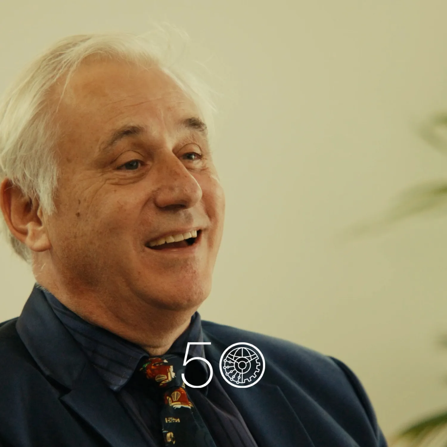 A person wearing a dark suit jacket, navy shirt, and patterned tie sits indoors against a light wall. Green plants frame the right side of the scene, and a “50” anniversary-style graphic appears at the bottom of the image.