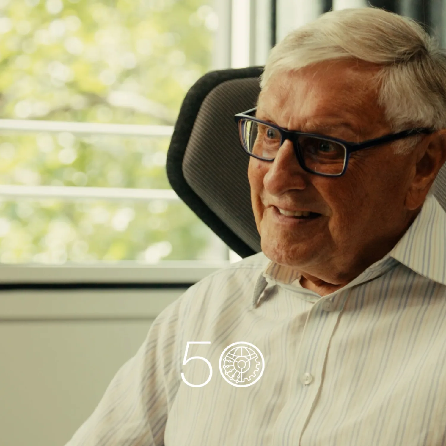 A person wearing a light striped button-up shirt sits in a cushioned chair beside a large window with green trees visible outside. Dark curtains frame part of the background, and a “50” anniversary-style graphic appears at the bottom of the image.