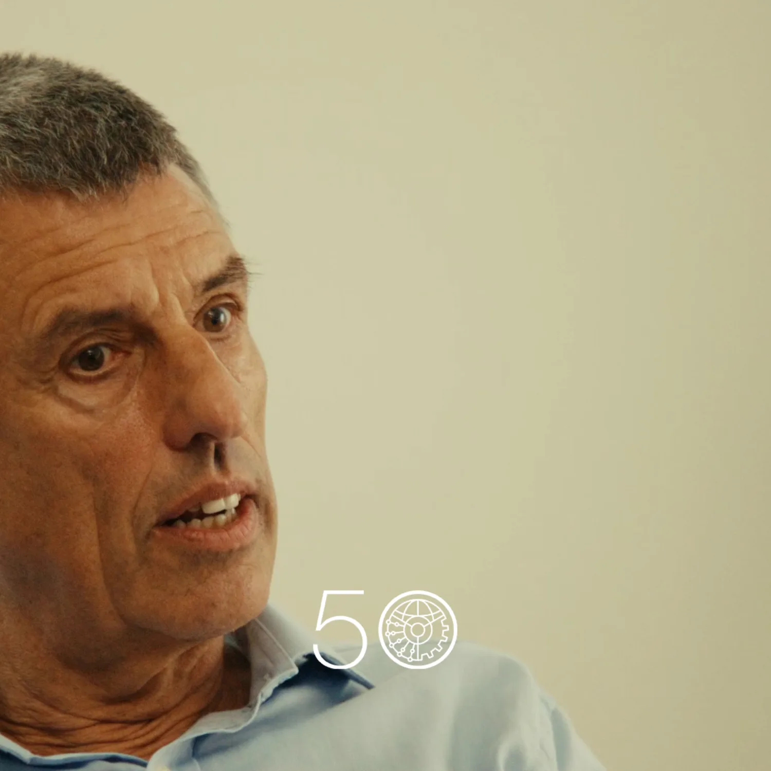 A person in a light blue collared shirt sits indoors against a plain light-colored wall. Part of a green plant is visible on the right, and a “50” anniversary-style graphic appears near the bottom of the image.