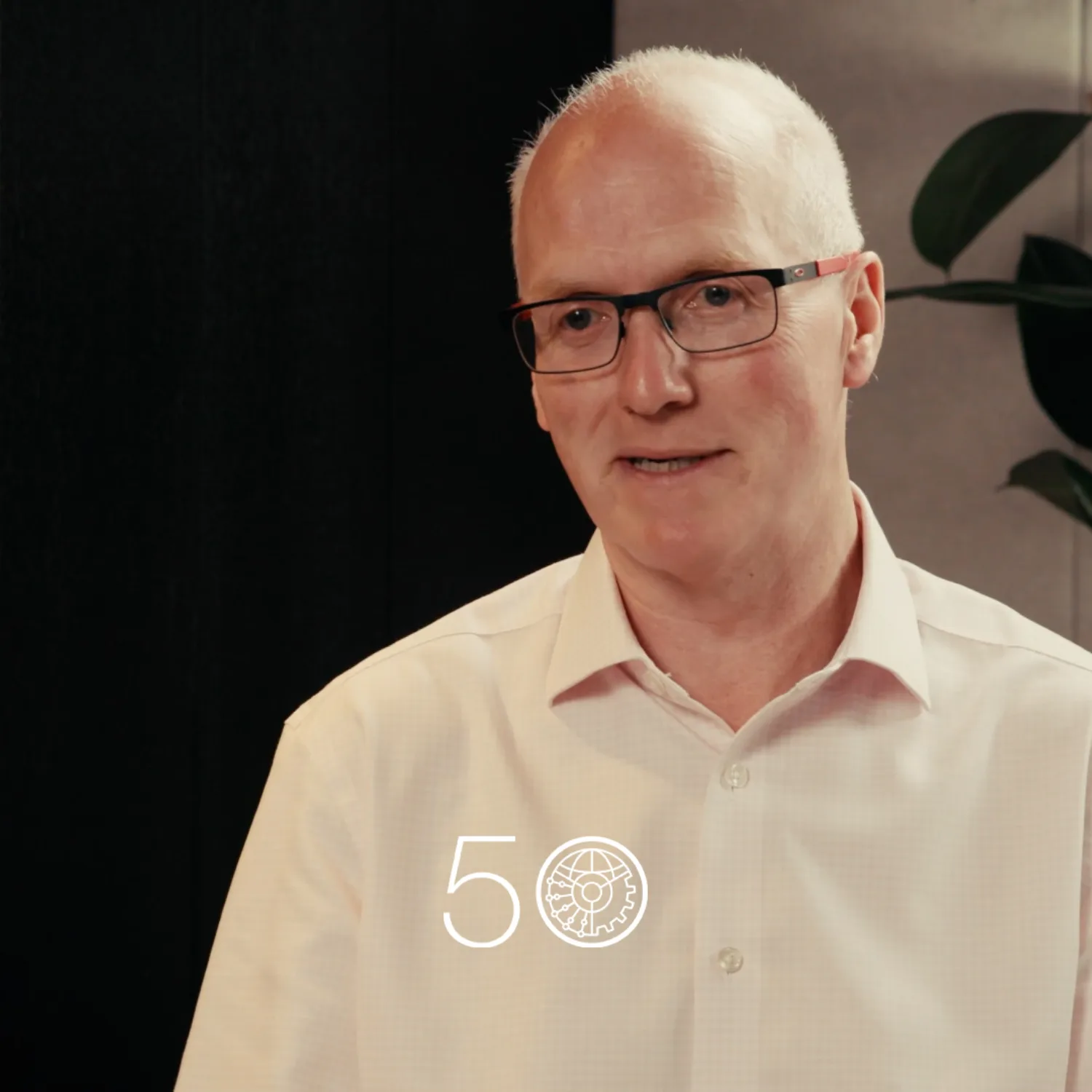 A person wearing a light pink button-up shirt sits indoors against a dark wall, with a tall green plant on the right. A “50” anniversary-style graphic is displayed near the bottom of the image.