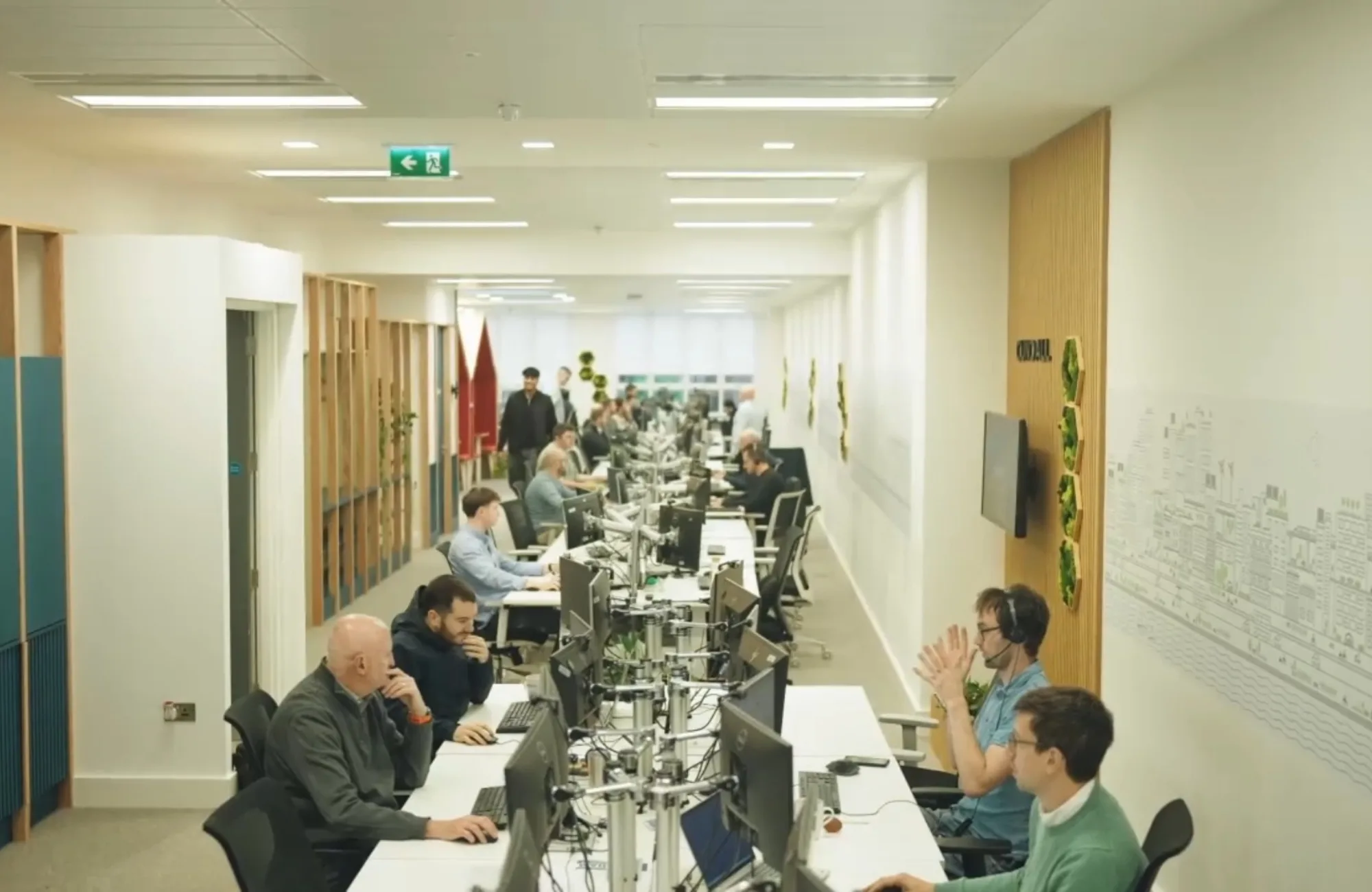 Open-plan office workspace with people seated at rows of desks and computer monitors beneath linear ceiling lights.