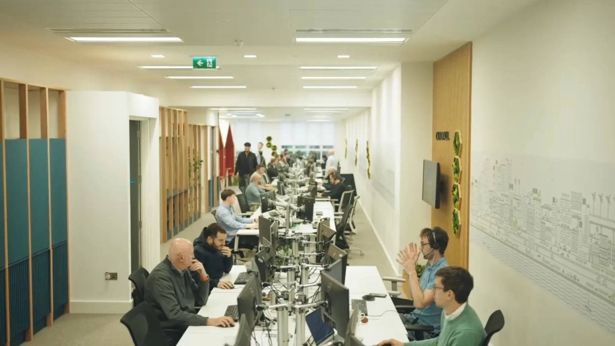 Open-plan office workspace with people seated at rows of desks and computer monitors beneath linear ceiling lights.