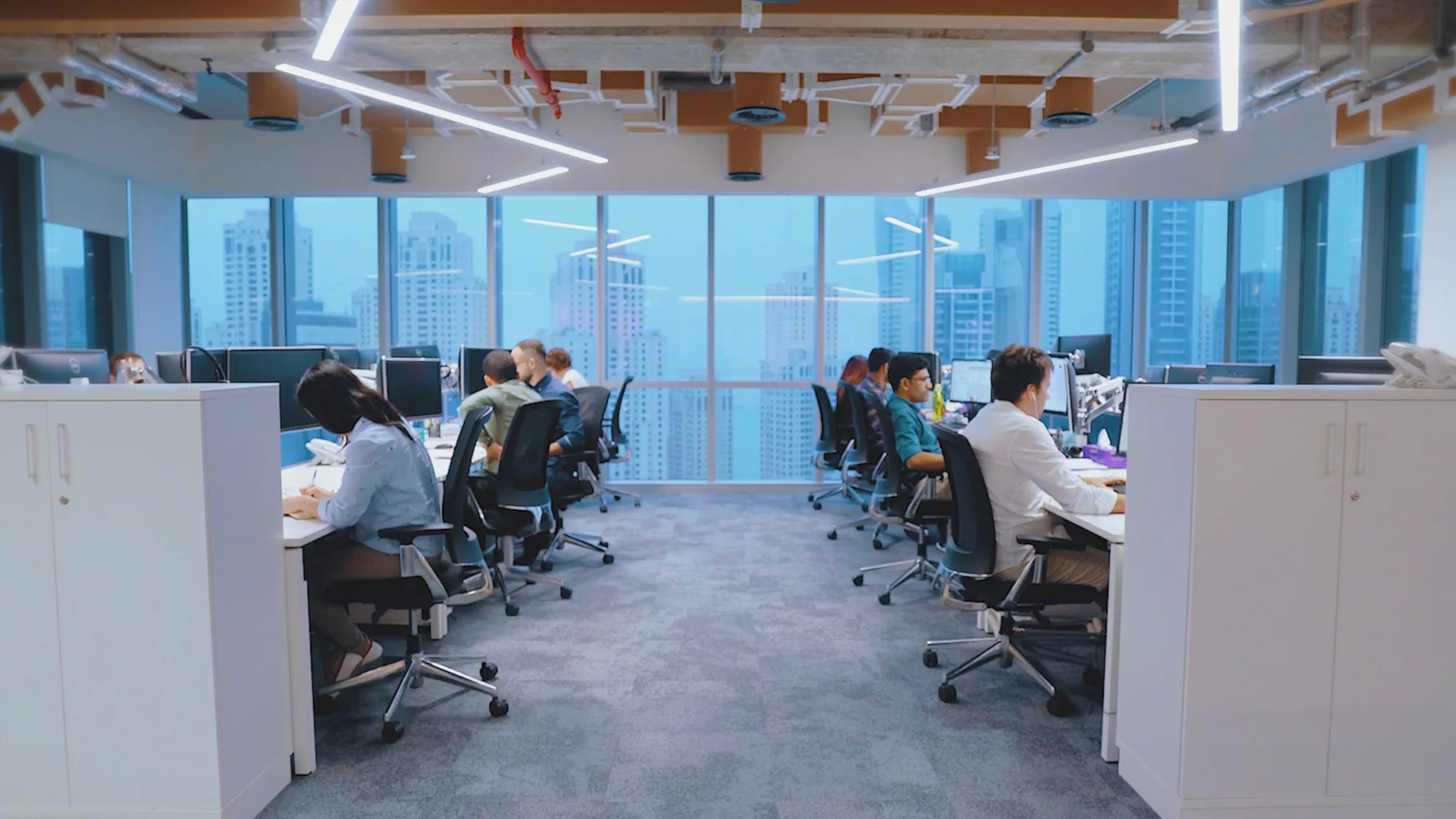 Two opposing rows of office workers at desk chairs in front of computers, in the background is a window looking out of a high rise building onto the Dubai skyline