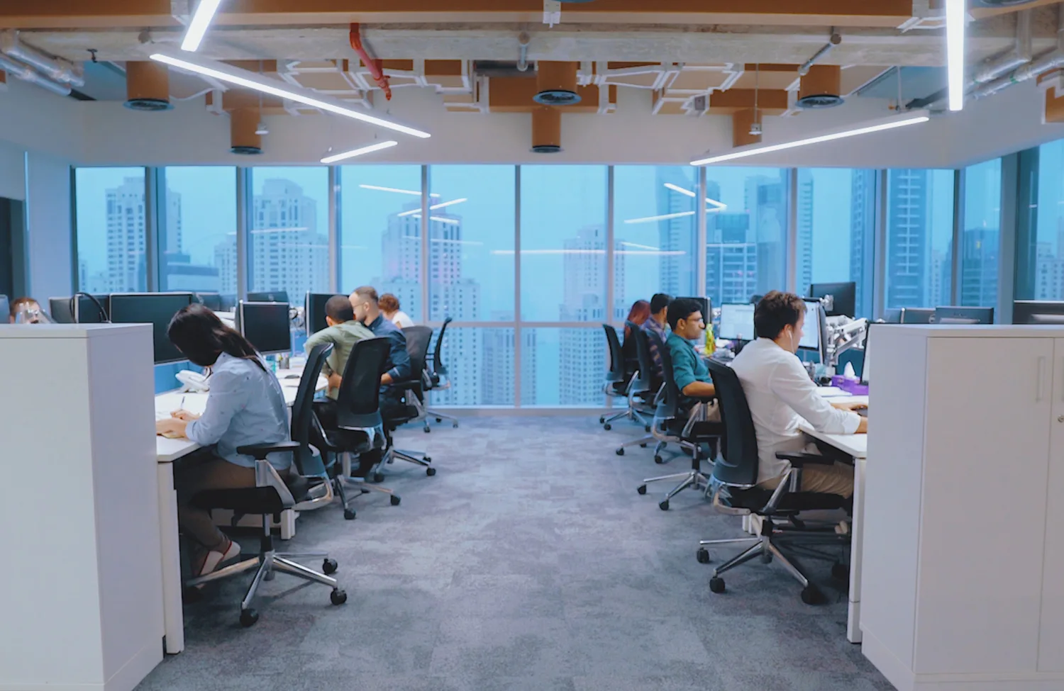 Two opposing rows of office workers at desk chairs in front of computers, in the background is a window looking out of a high rise building onto the Dubai skyline
