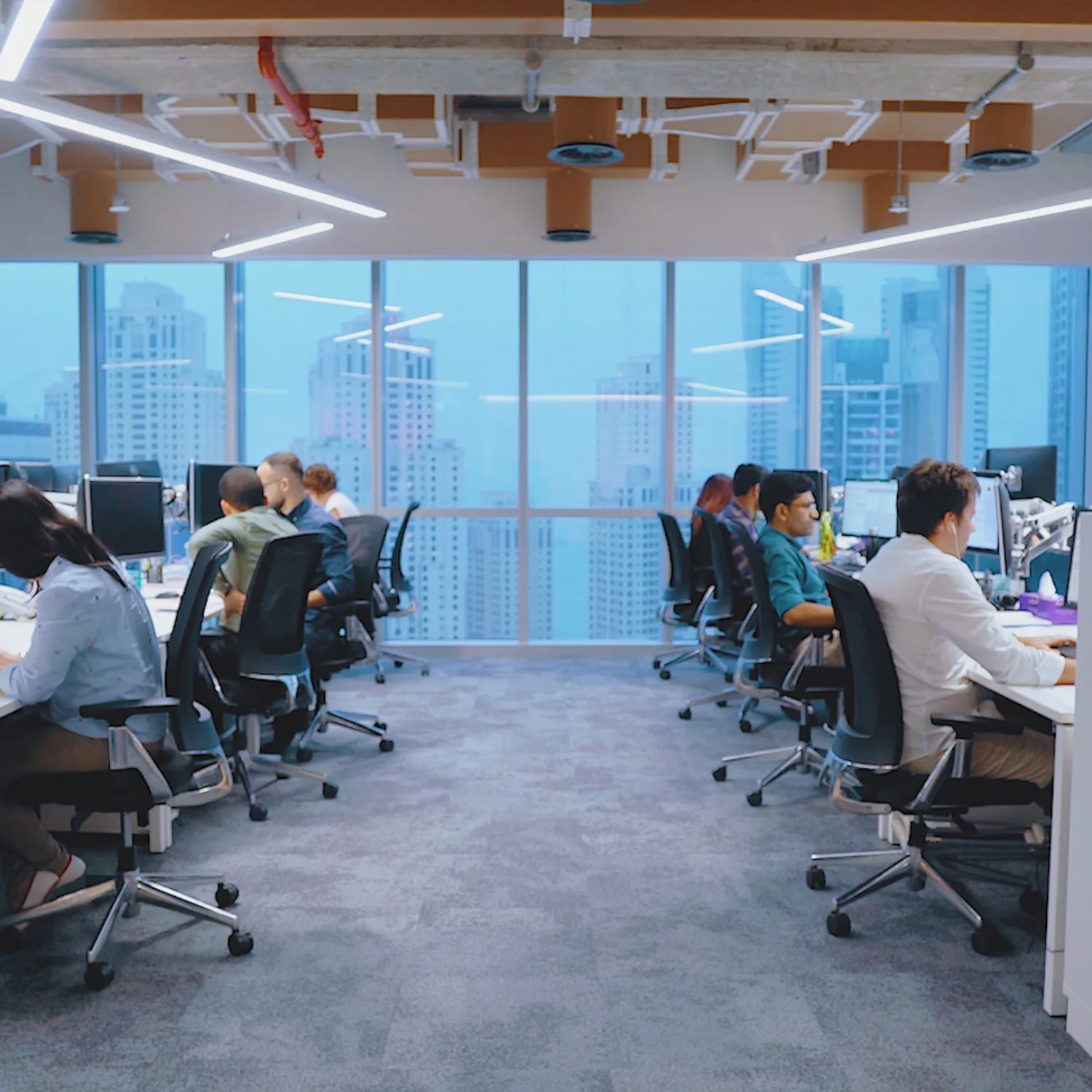 Two opposing rows of office workers at desk chairs in front of computers, in the background is a window looking out of a high rise building onto the Dubai skyline