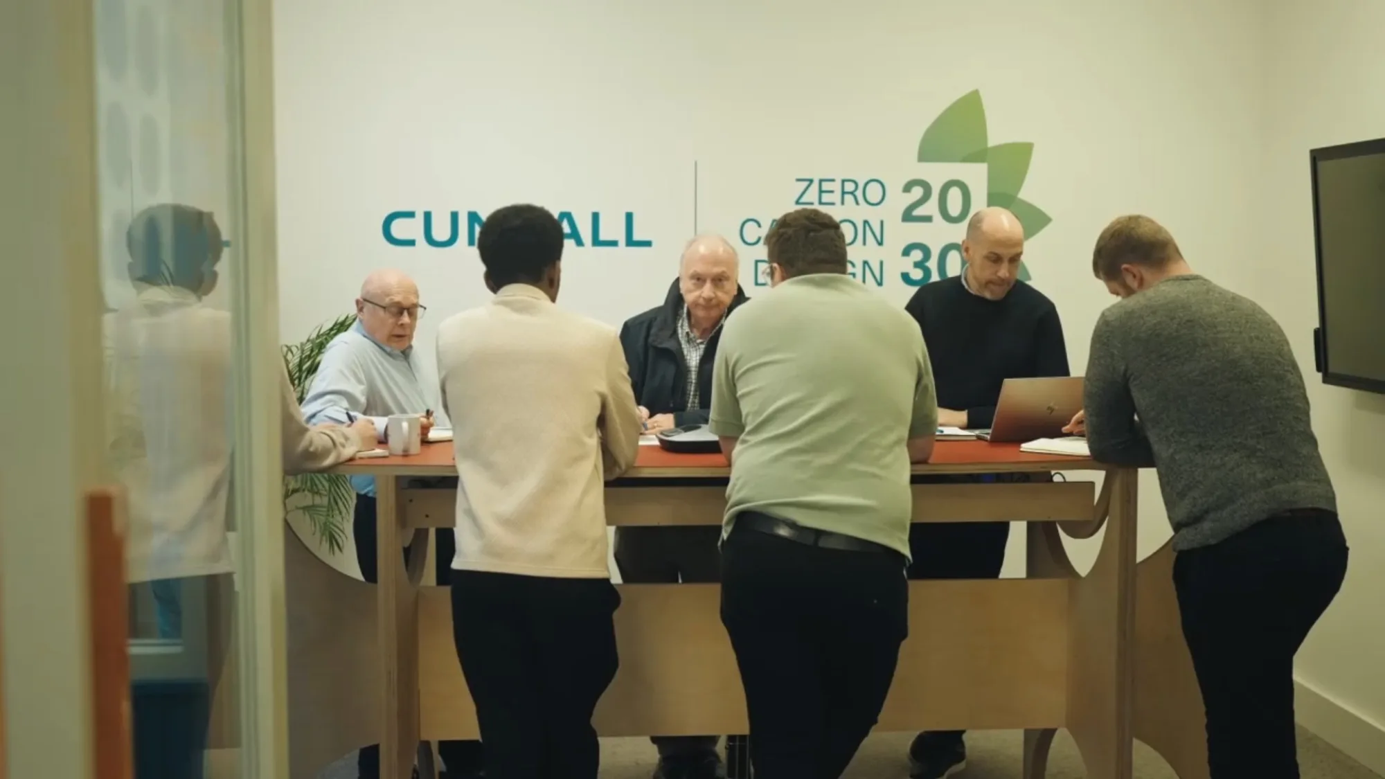 People gathered around a high table with a laptop in a glass-walled workspace featuring Zero Carbon Design 2030 graphics.