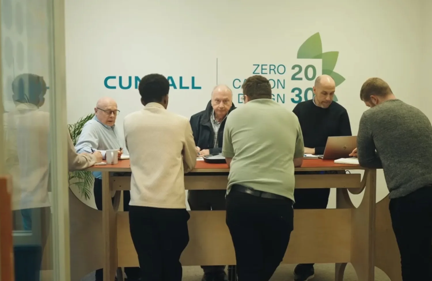 People gathered around a high table with a laptop in a glass-walled workspace featuring Zero Carbon Design 2030 graphics.