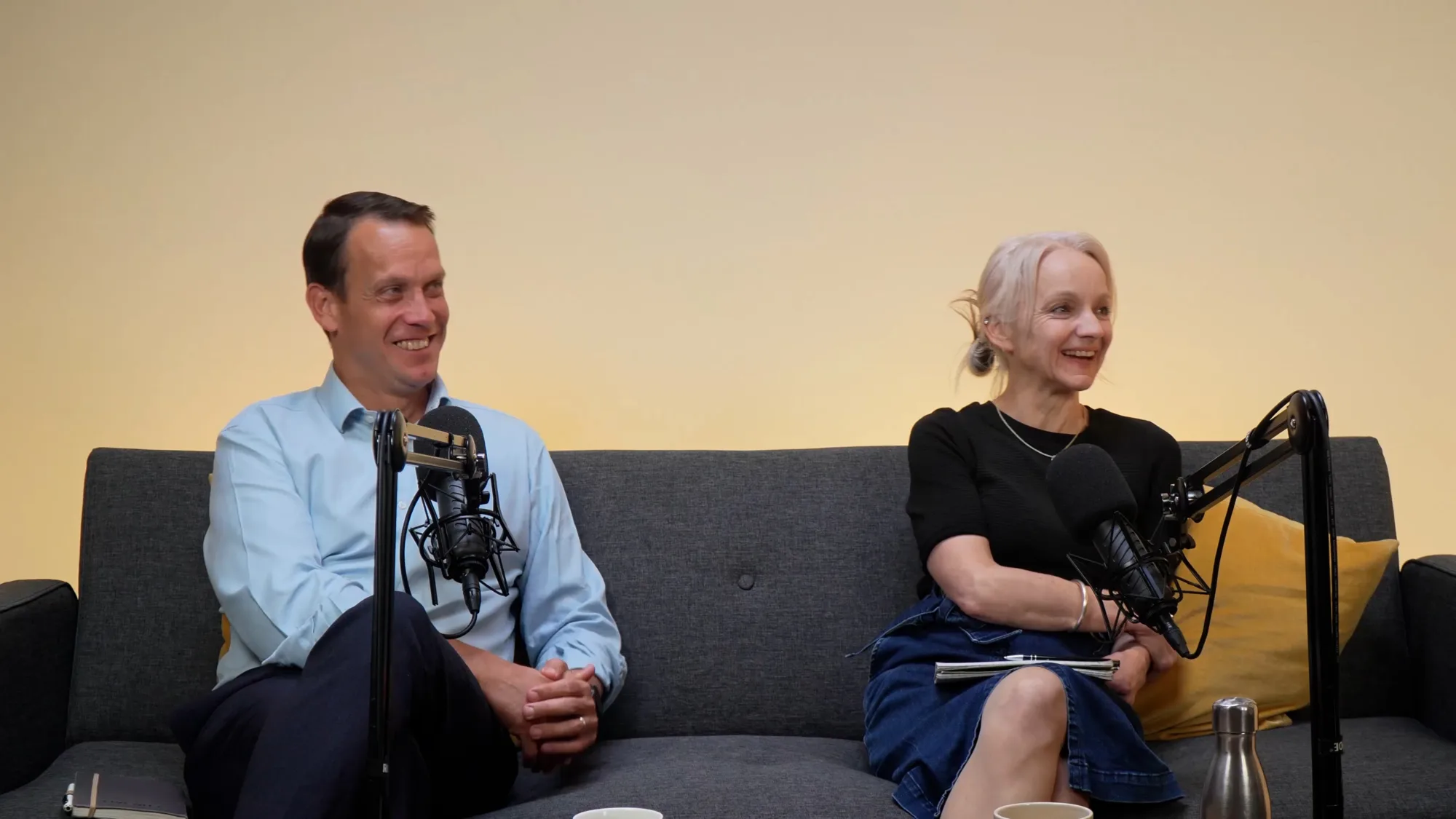 Robin and anna sat on a sofa with podcast mics