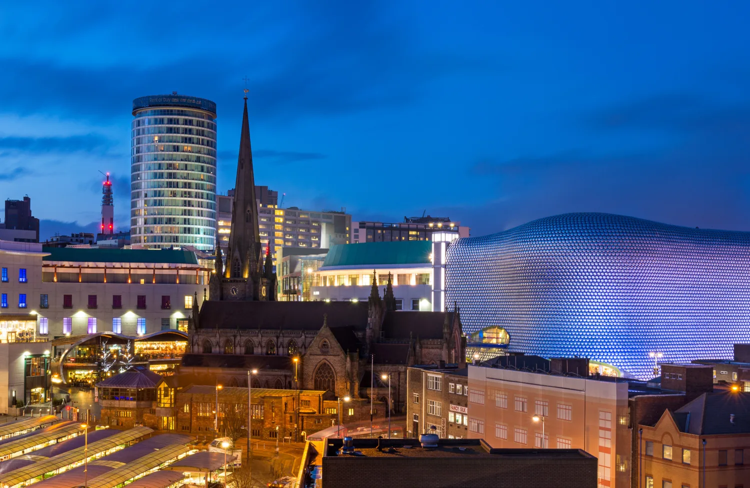 Birmingham city, a church in front of a cylindrical building and shopping centre with lit streets against a dark blue sky