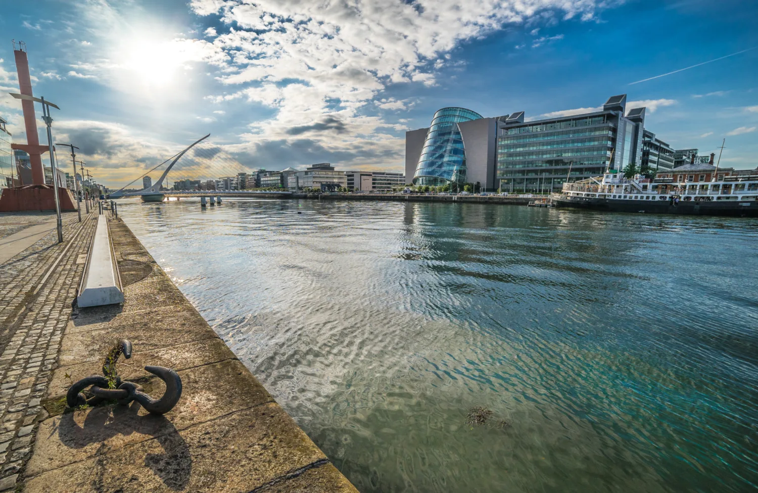 River and paved embankment with pedestrian bridge and a sunlit blue sky