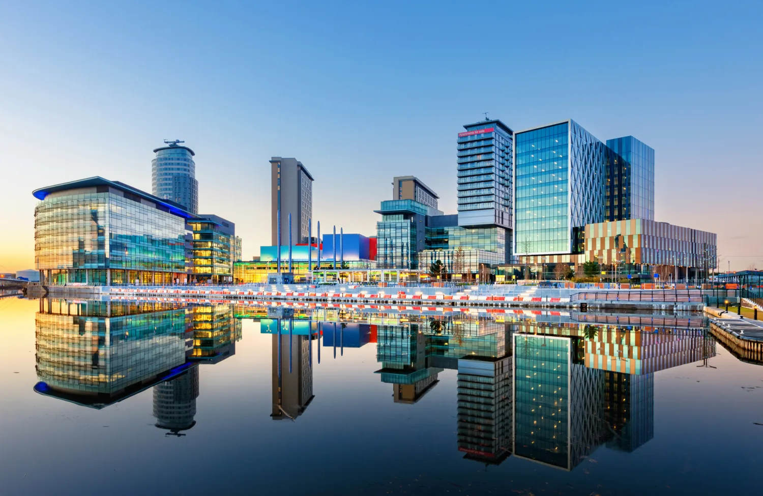 Lit modern buildings reflected in still water, dusk sky
