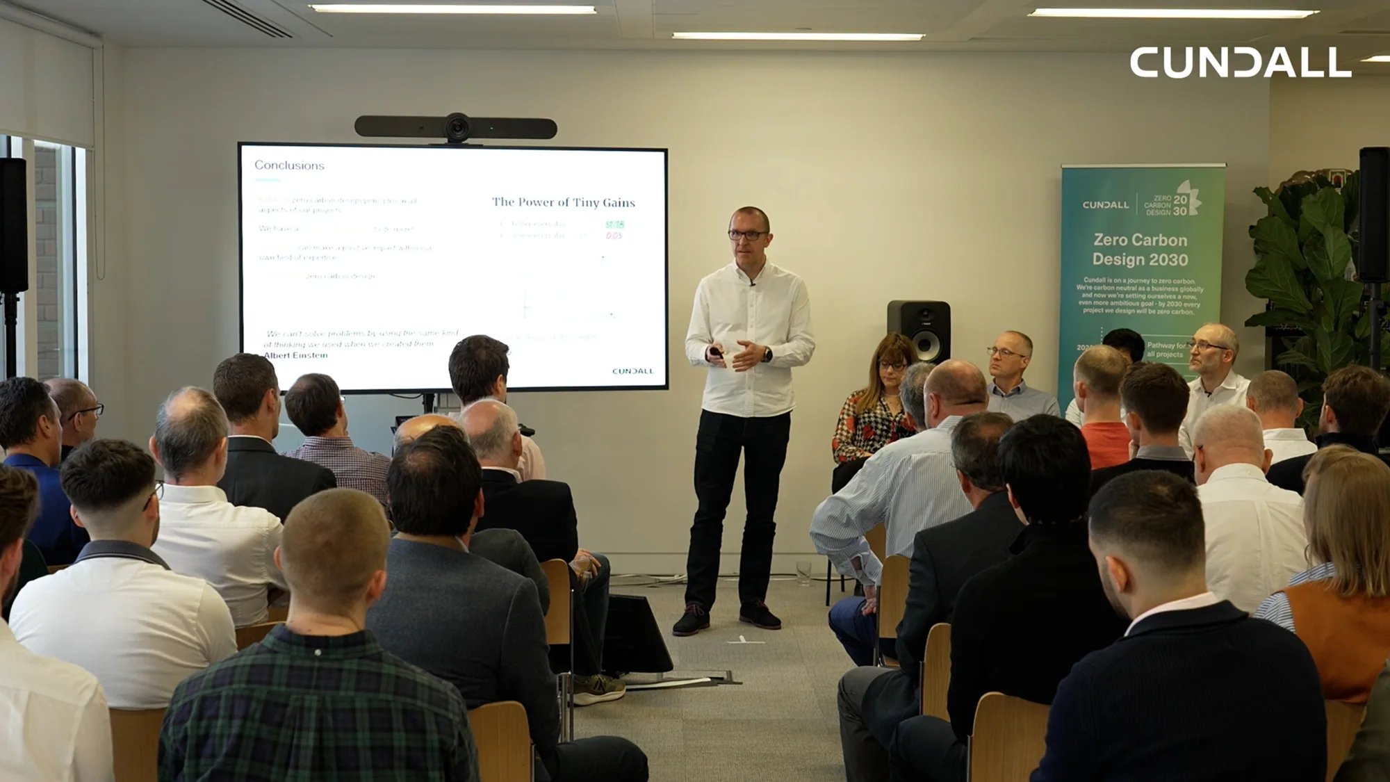 Peter Liddle presenting standing in front of the screen with room of people watching