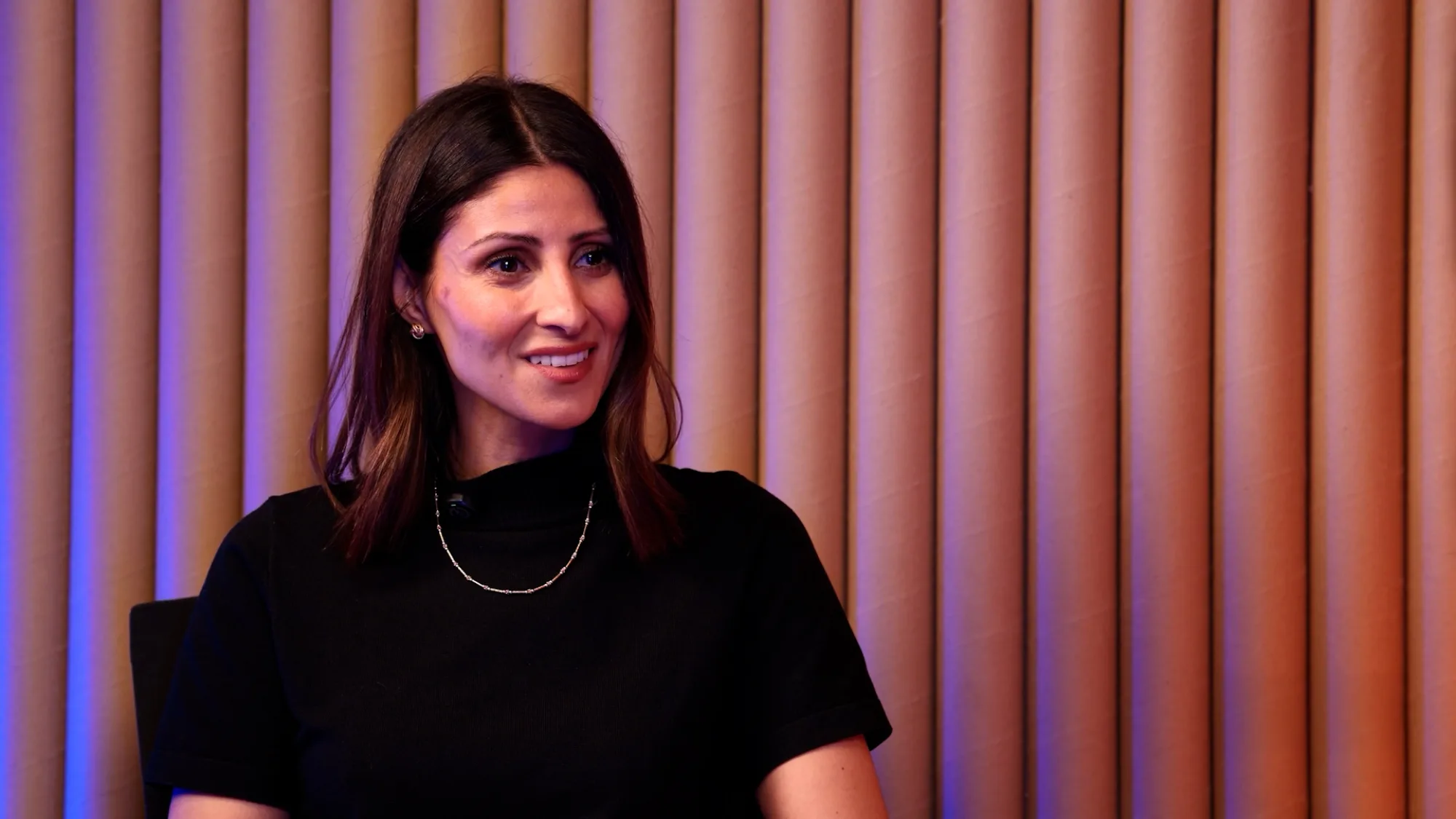 Noelle Halabi, with black long hair and black dress, being interviewed and smiling.
